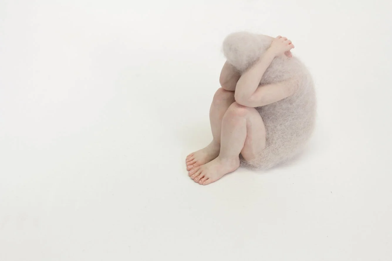 A felted figurine is sitting with knees up and arms over their head in a minimalist setting with a white background.