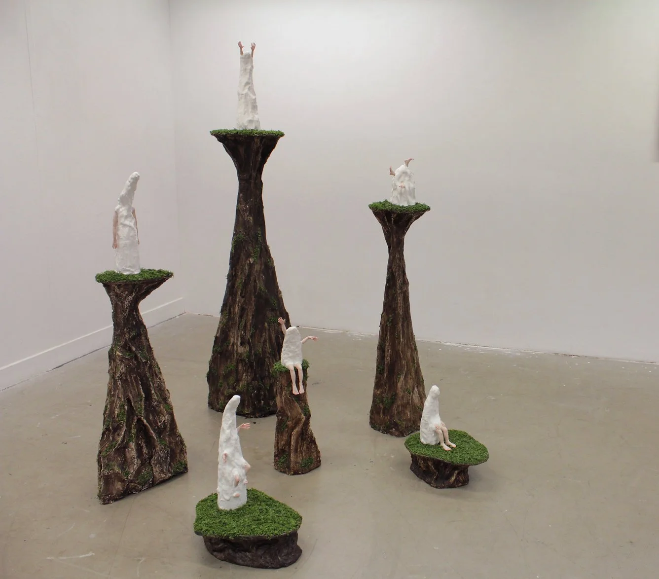 Art installation with tree bark-like pedestals topped with small white sculptures of human figures, some with extended arms or sitting, against a plain white gallery wall.