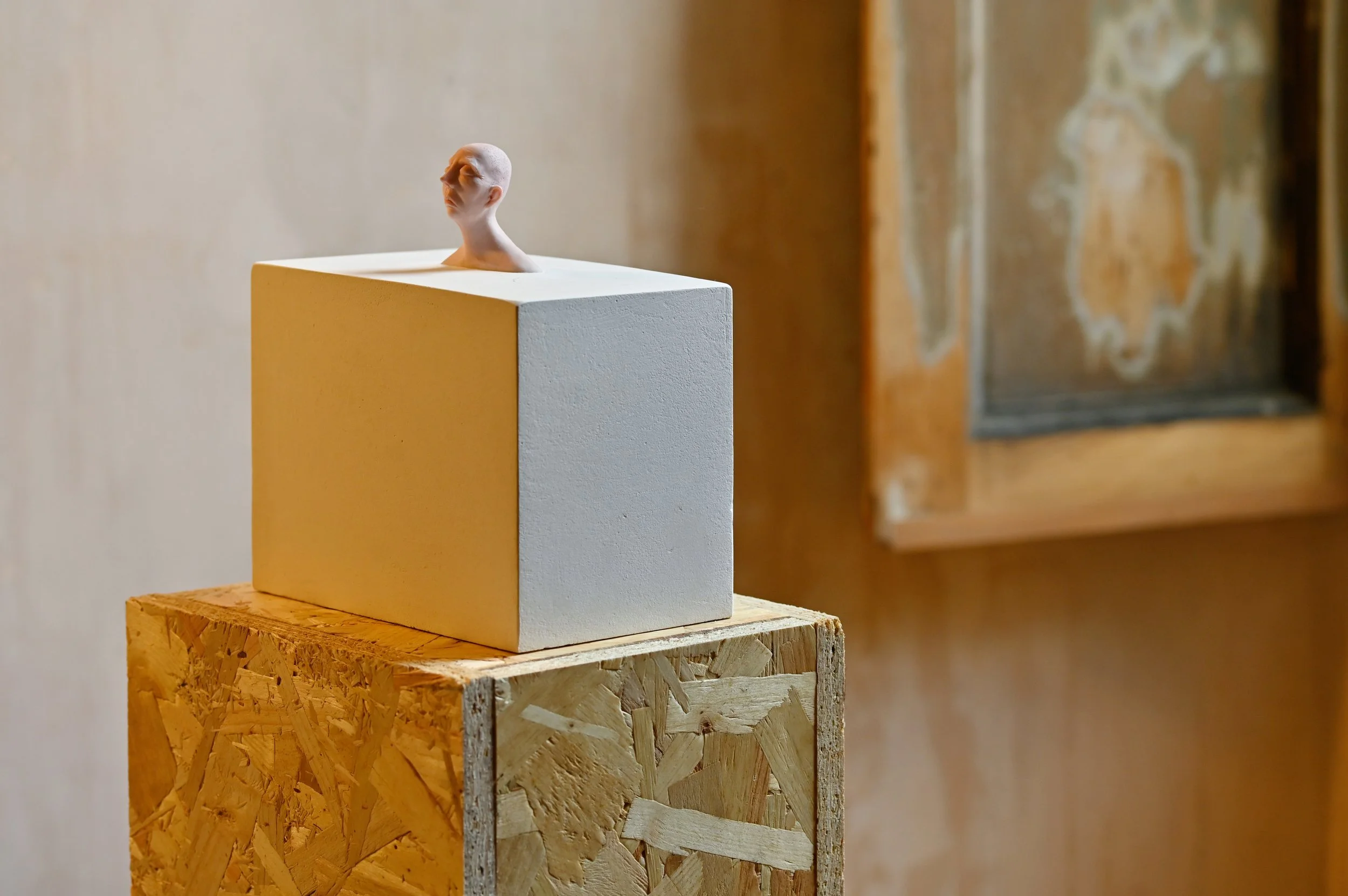 A small sculpture of a human head with closed eyes, emerging from a white cube placed on an OSB wood pedestal, with a blurred picture frame in the background.