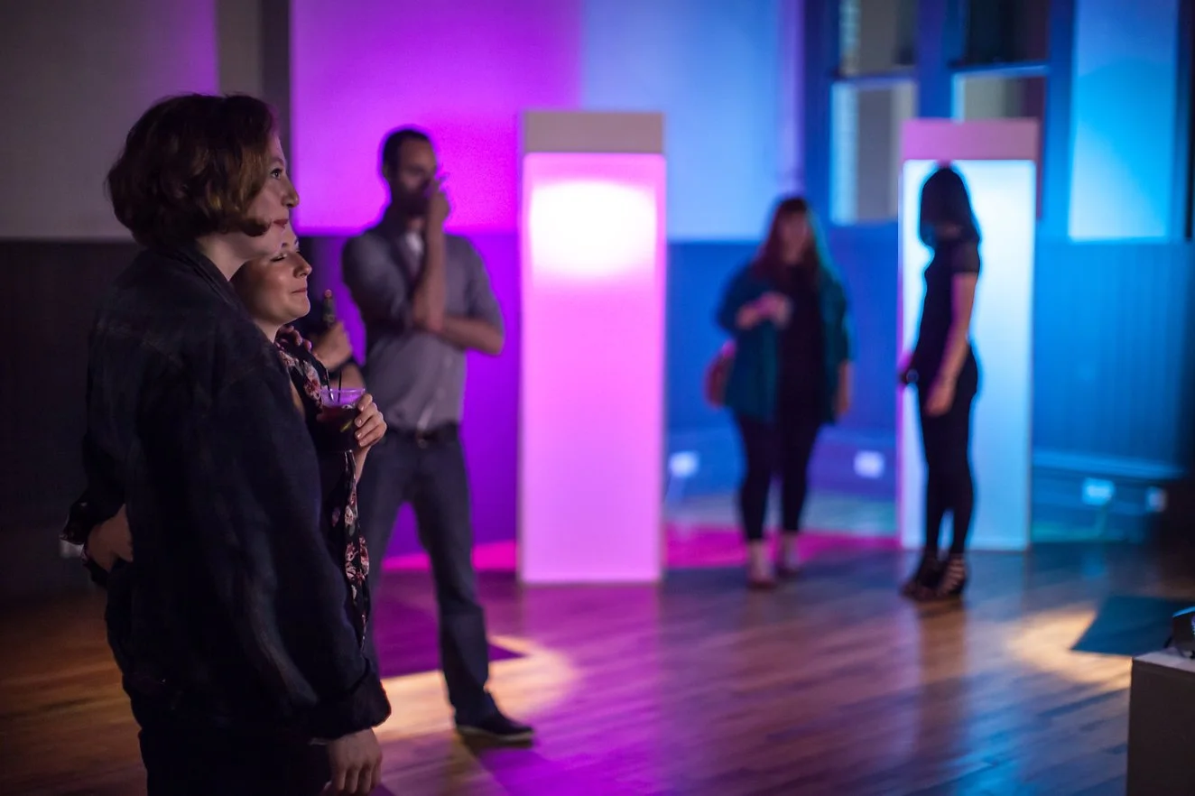 People standing in a dimly lit room with colorful purple and blue lighting, some holding drinks, engaged in conversation or observing surroundings.