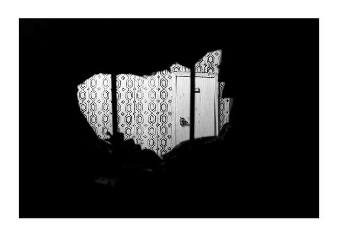 View through a hole in a wall showing a patterned wallpaper and a door with a handle.