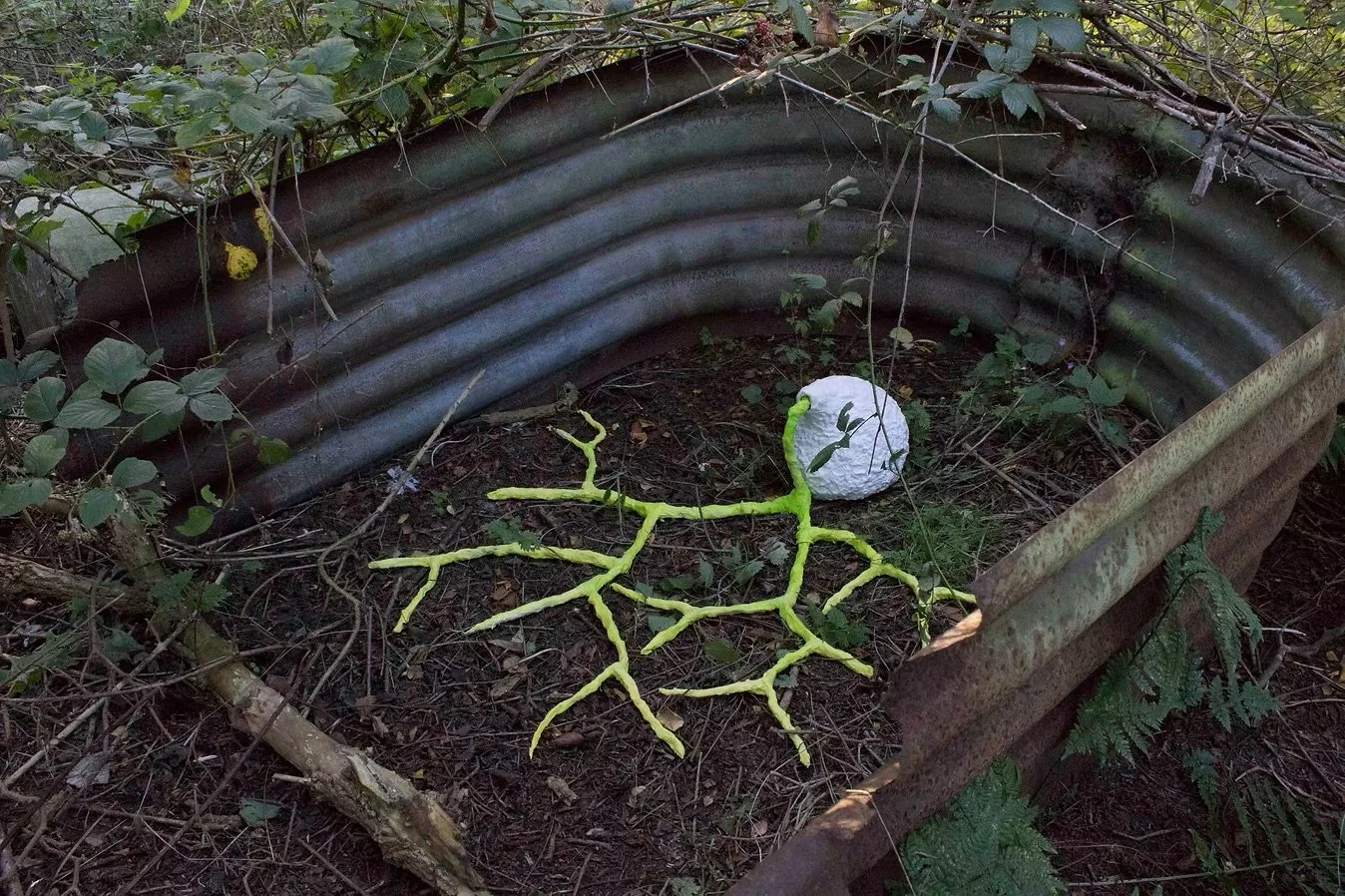 A small garden scene with a corrugated metal border, some plants and leaves, a white sculpture has twisted green vines emerging from it on to  the soil.