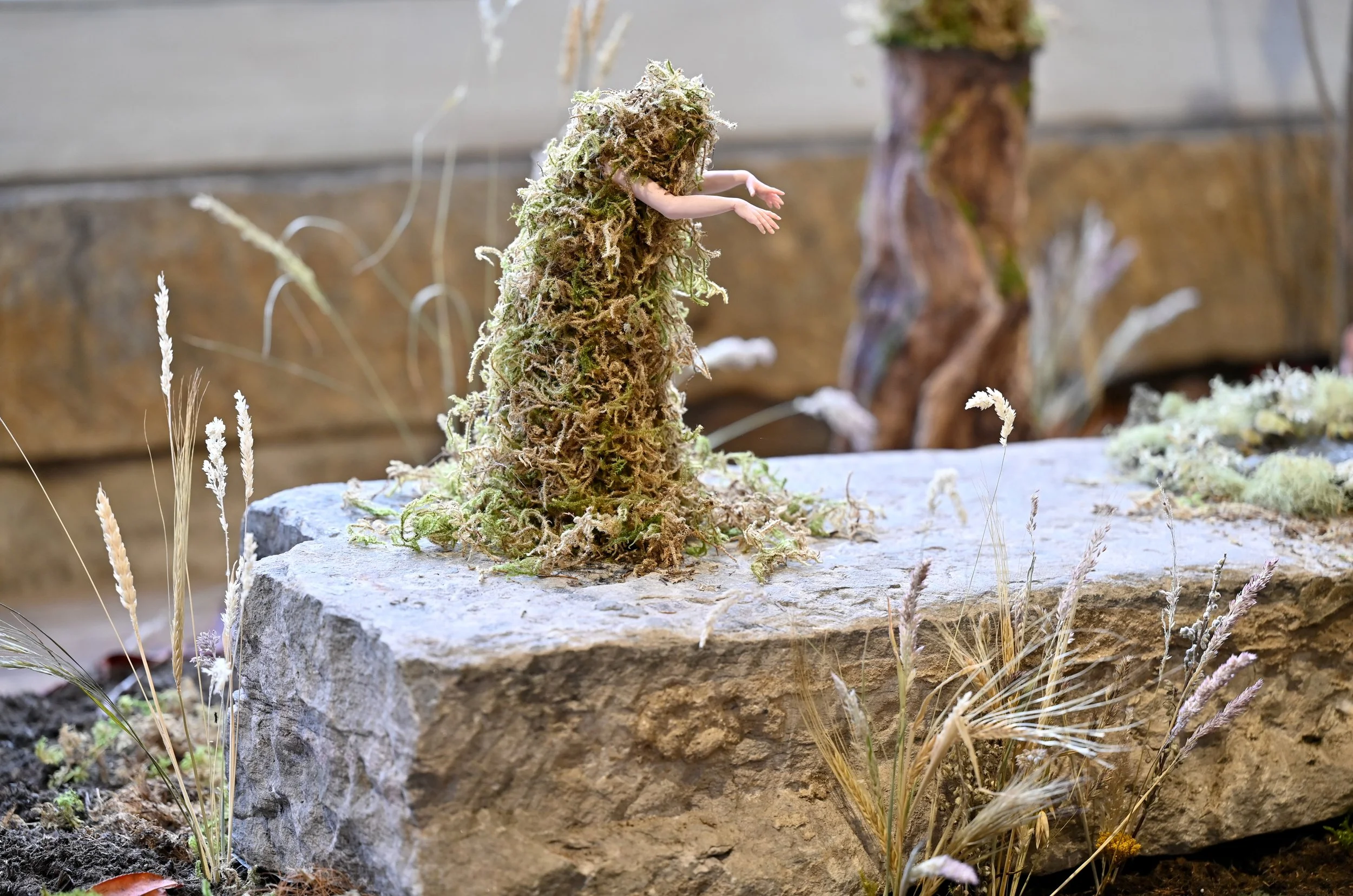 A small figure with human arms is emerging from a large, bushy, green mossy mound on a stone surface in a garden setting with plants and rocks around.