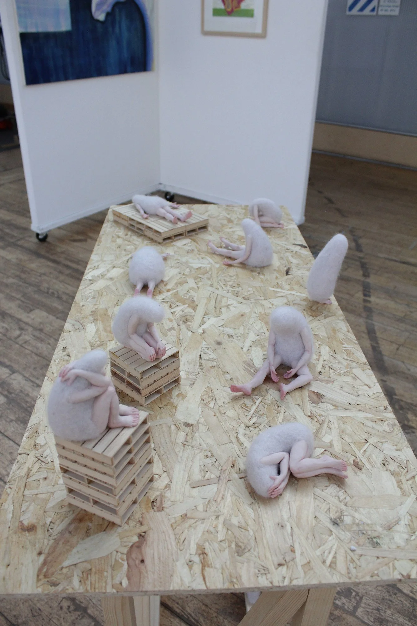 Felted sculptures with human-like limbs, posed on stacked wooden pallets and a wooden table in an art display.