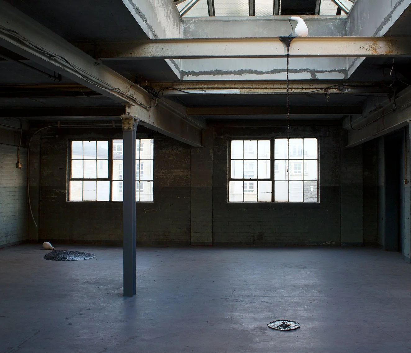 Empty industrial space with large windows, exposed metal beams, and a skylight roof, featuring a metal pole and a small light fixture on the floor.
