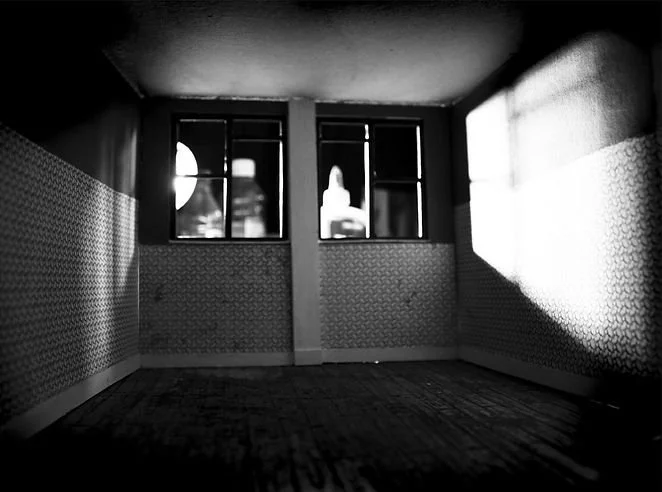 Empty room with patterned wallpaper, wooden floor, and three windows with sunlight streaming through.