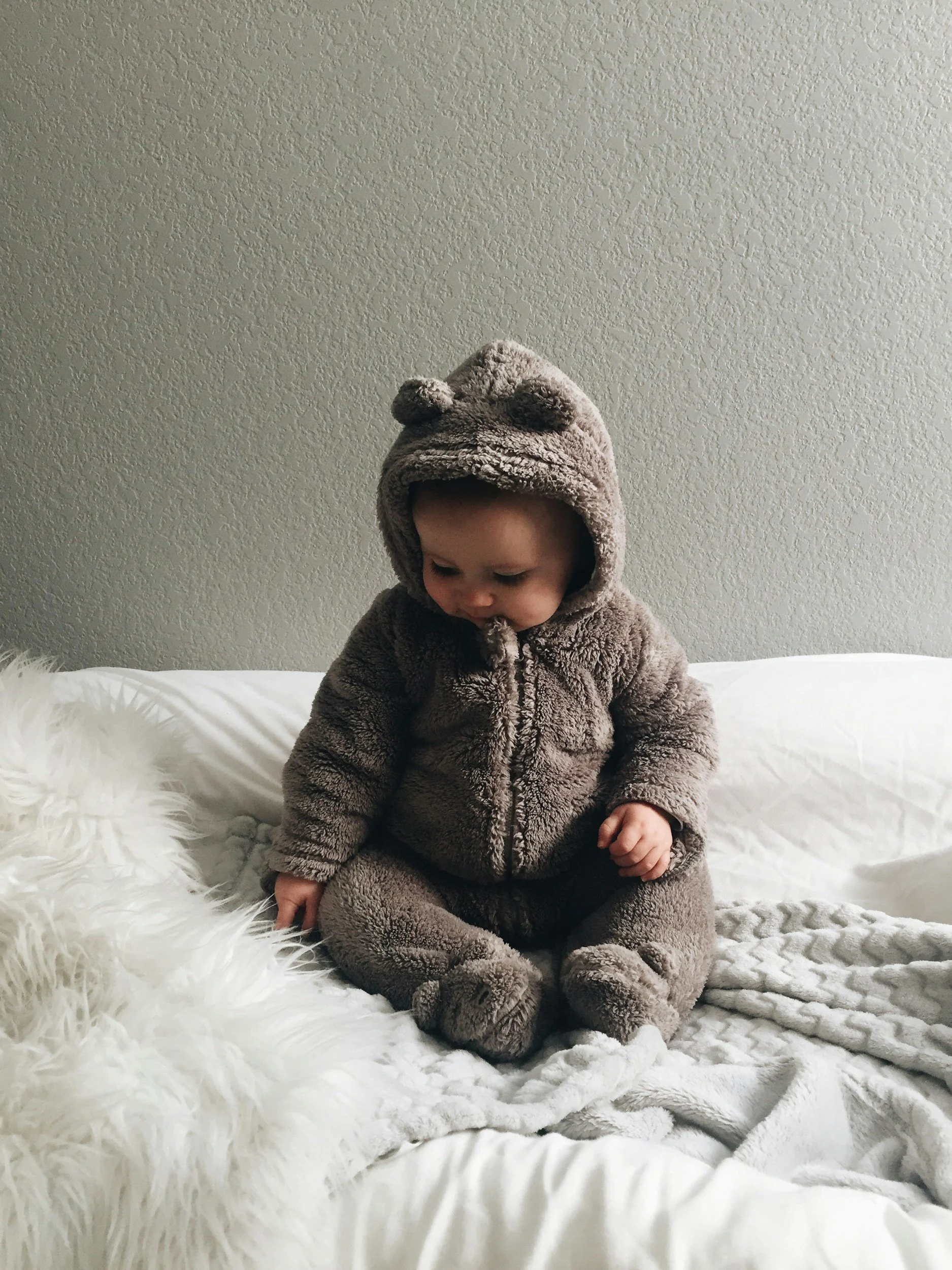 A baby dressed in a plush, bear-themed hooded onesie with bear paw details, sitting on a white bed with a furry pillow and blanket, looking down.