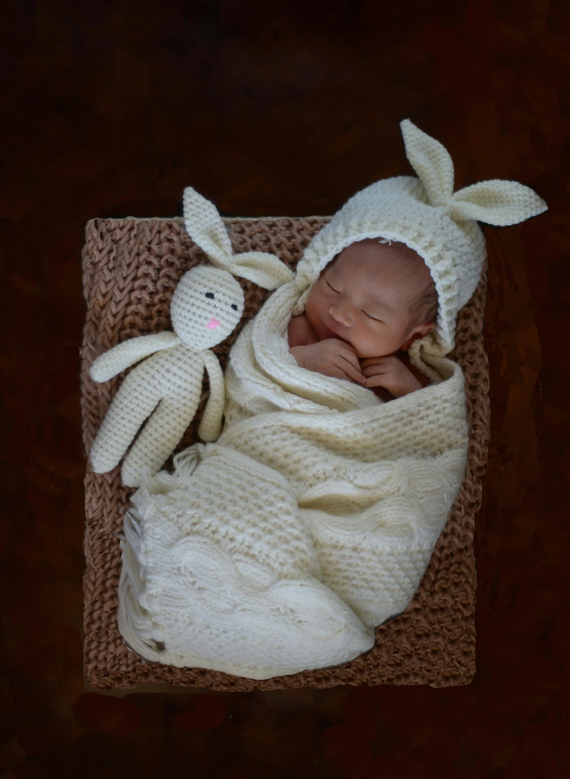 A newborn baby dressed in a knit bunny hat and blanket, lying on a textured brown blanket with a crocheted bunny plush toy beside them.