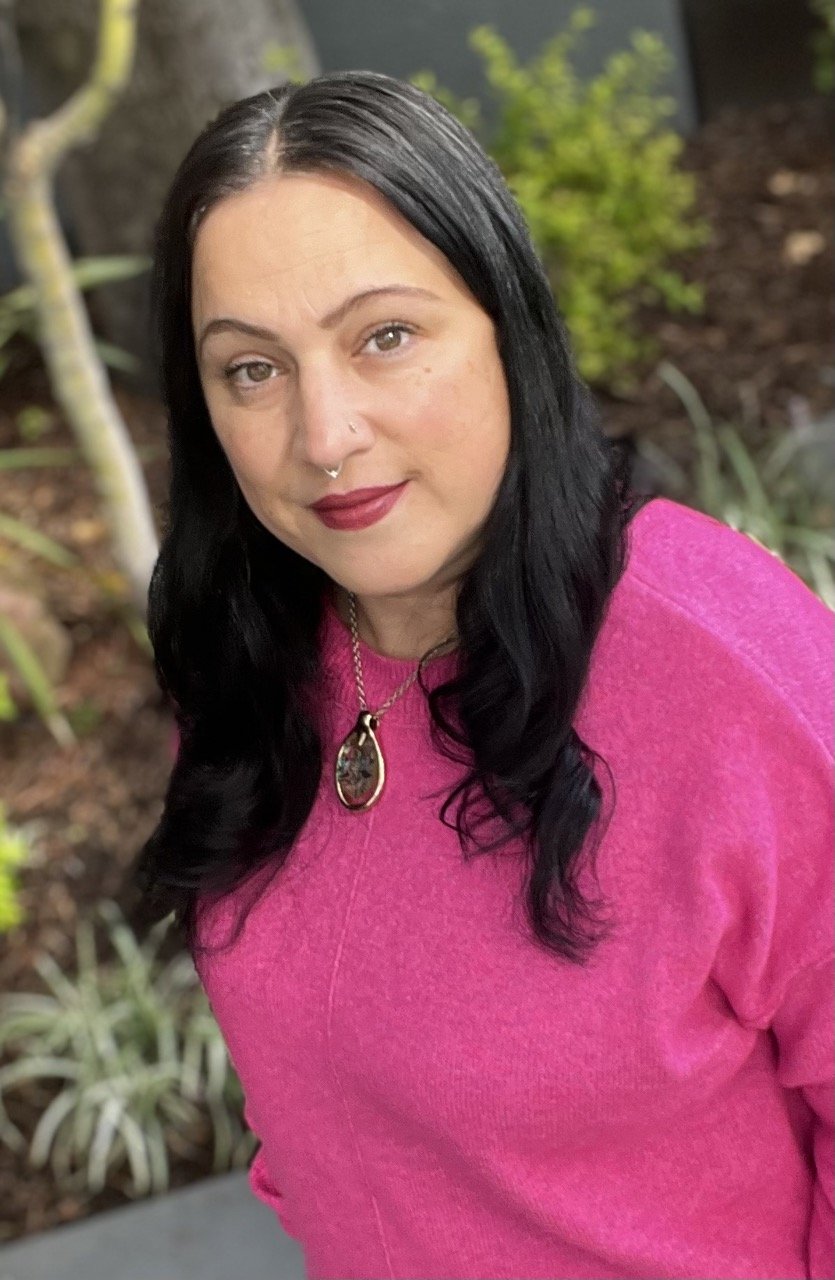 A woman with long black hair, wearing a pink sweater and red lipstick, standing outdoors near plants and bushes.