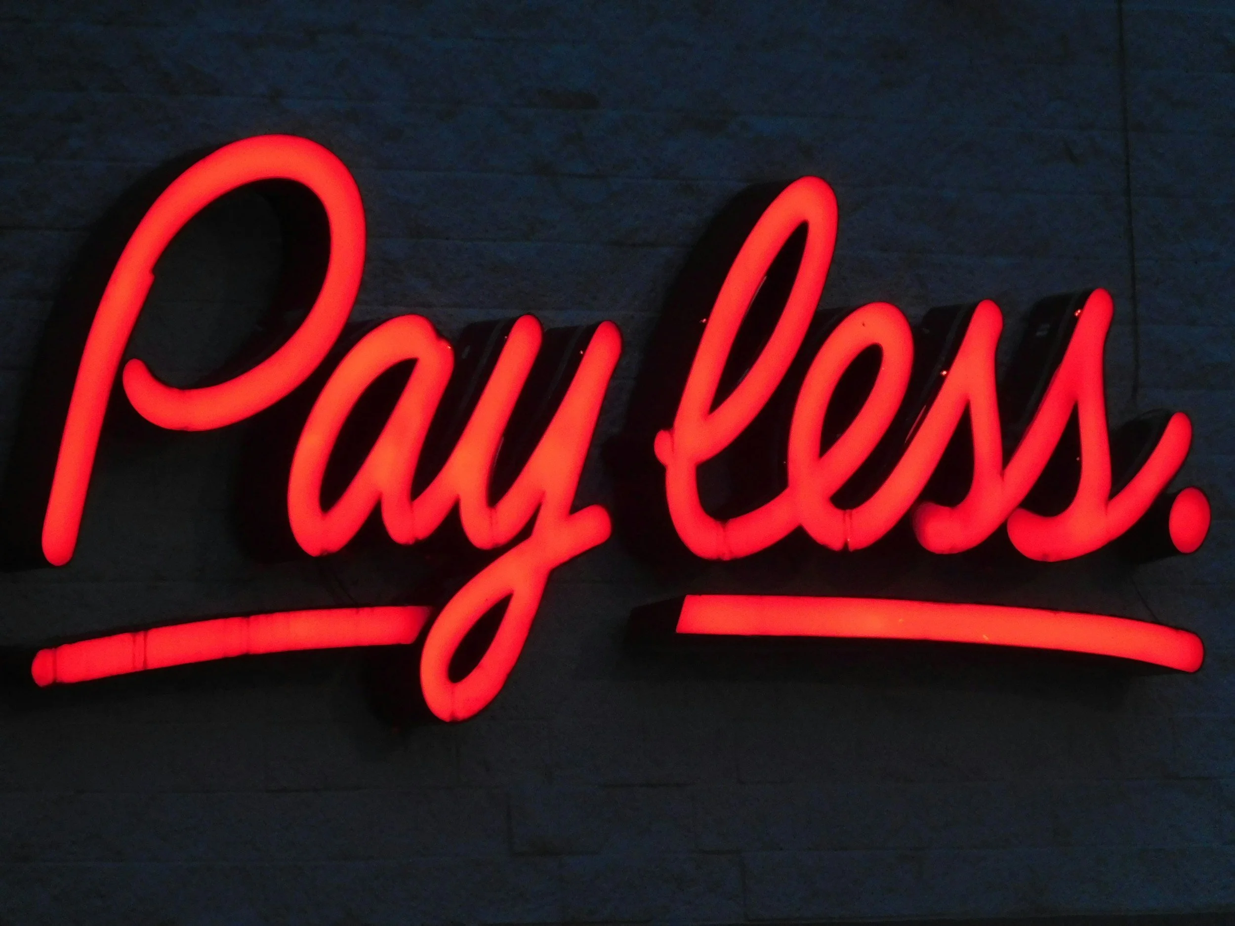 Red neon sign spelling 'Payless' on a dark wall.