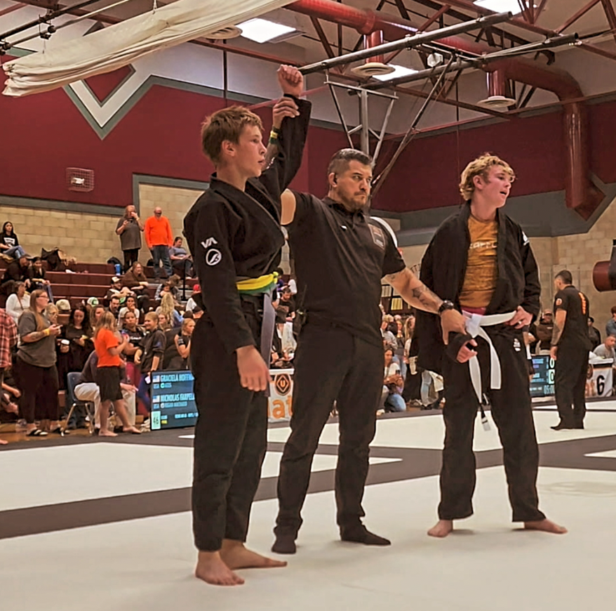 A female Brazilian Jiu-Jitsu competitor in a black gi with a yellow belt has her hand raised by the referee in an indoor gymnasium, indicating she is the winner of the match. Another competitor in a black gi and white belt stands nearby, with spectators watching in the background.