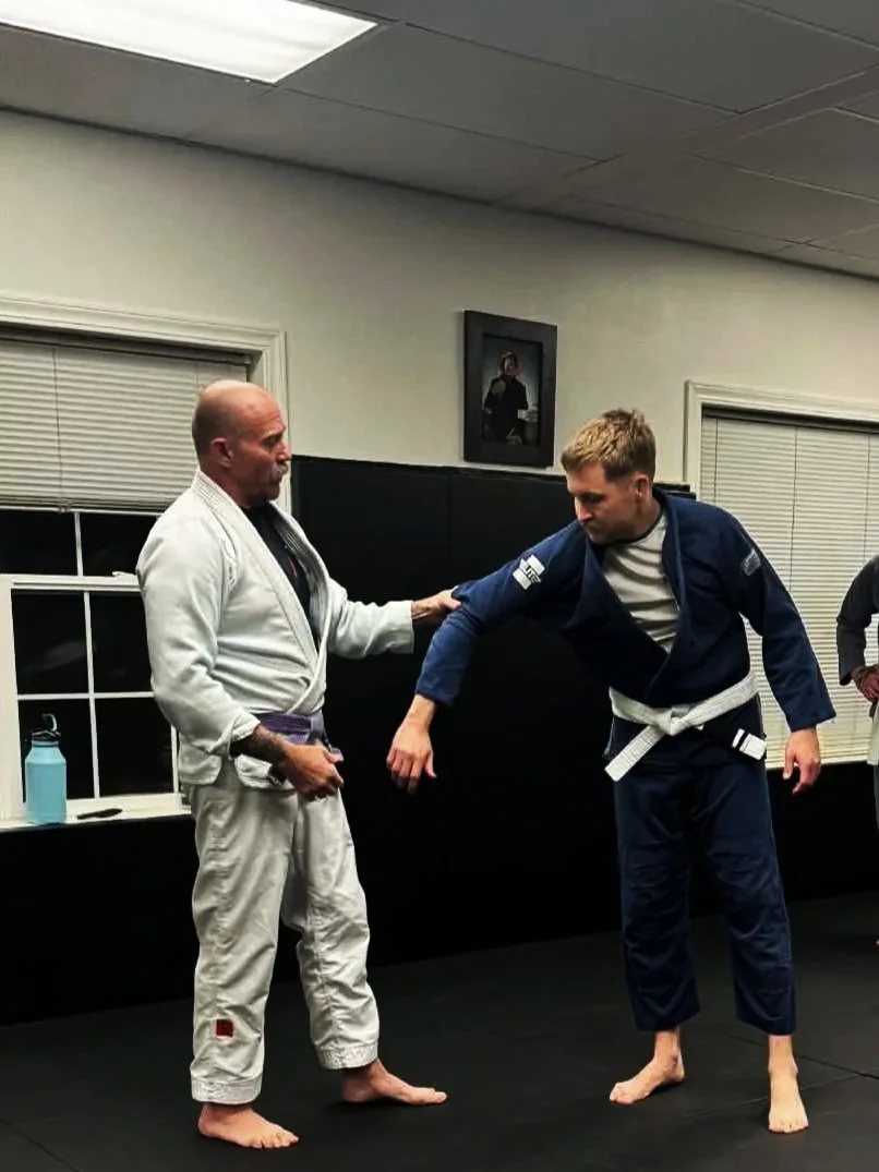 Two men practicing Brazilian Jiu-Jitsu on a black mat in a training room, one in a white gi and the other in a blue gi, with a portrait on the wall and a water bottle on a table in the background.