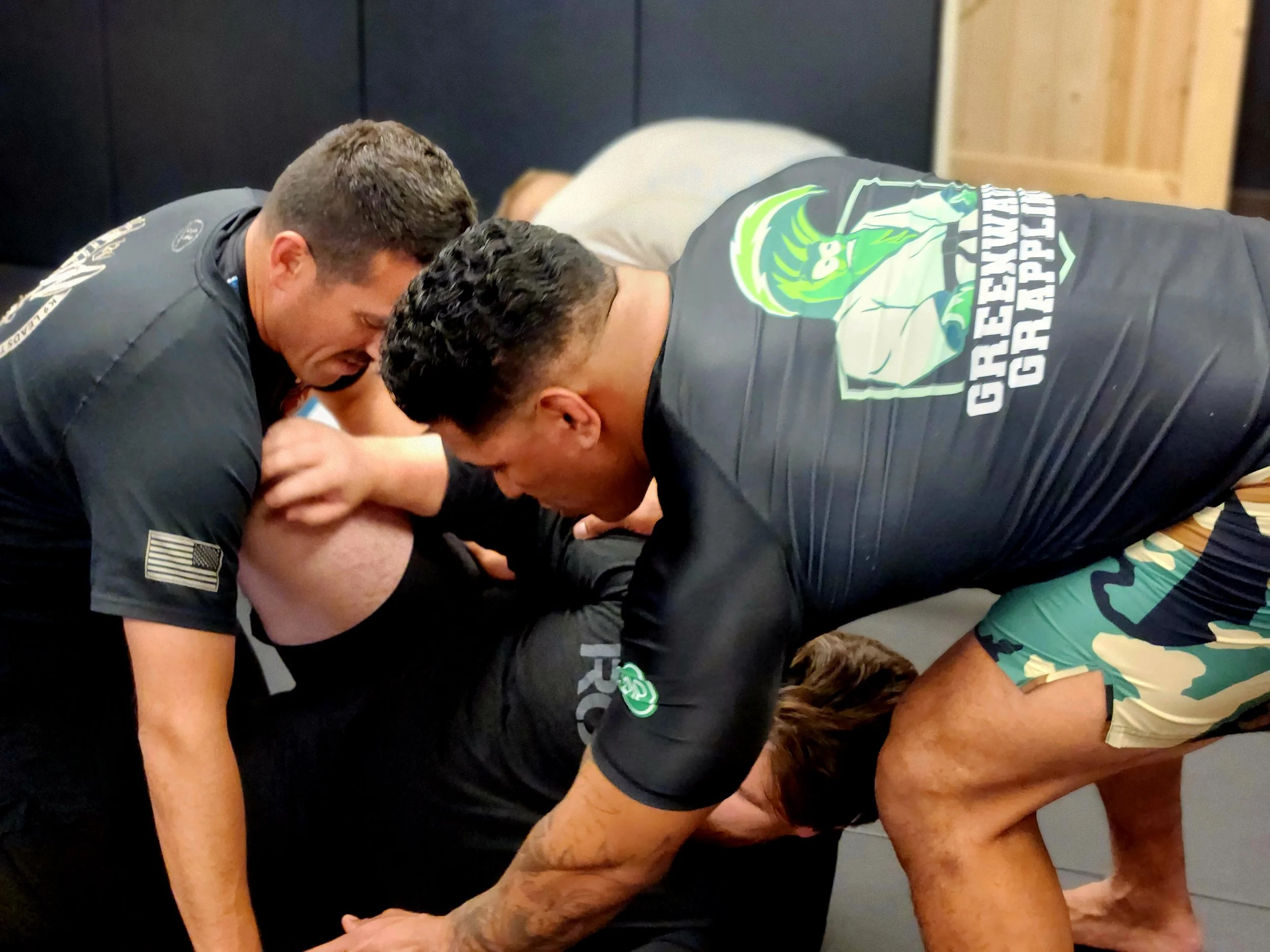 Two men in black T-shirts are grappling on the ground, wrestling or training, with one person pinning the other. They are in a room with dark walls and a wooden door.