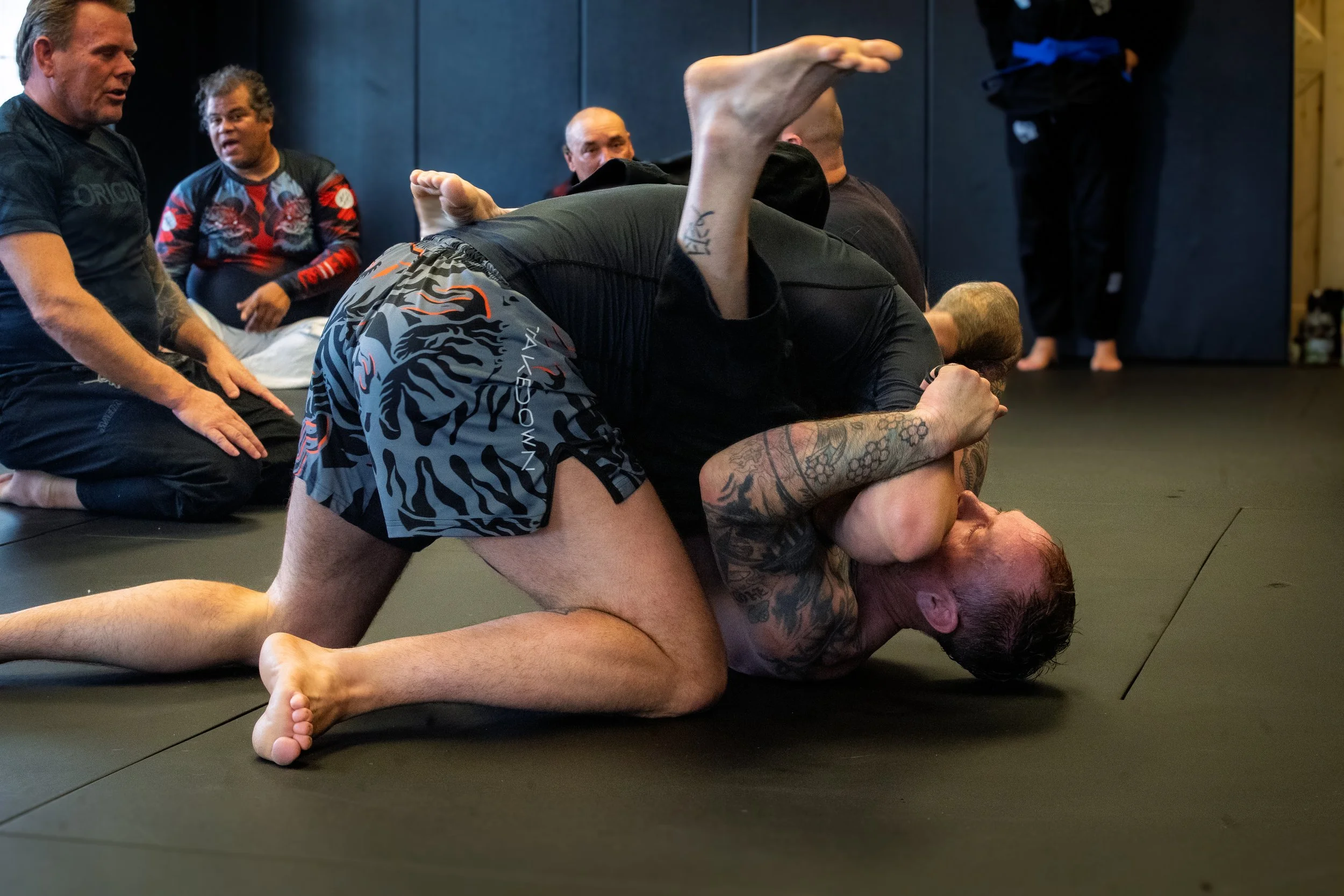 Two men practicing Brazilian Jiu-Jitsu on a black mat surrounded by other practitioners. One man is on top, pinning the other, who is on the ground with tattoos, in a grappling position during a training session.