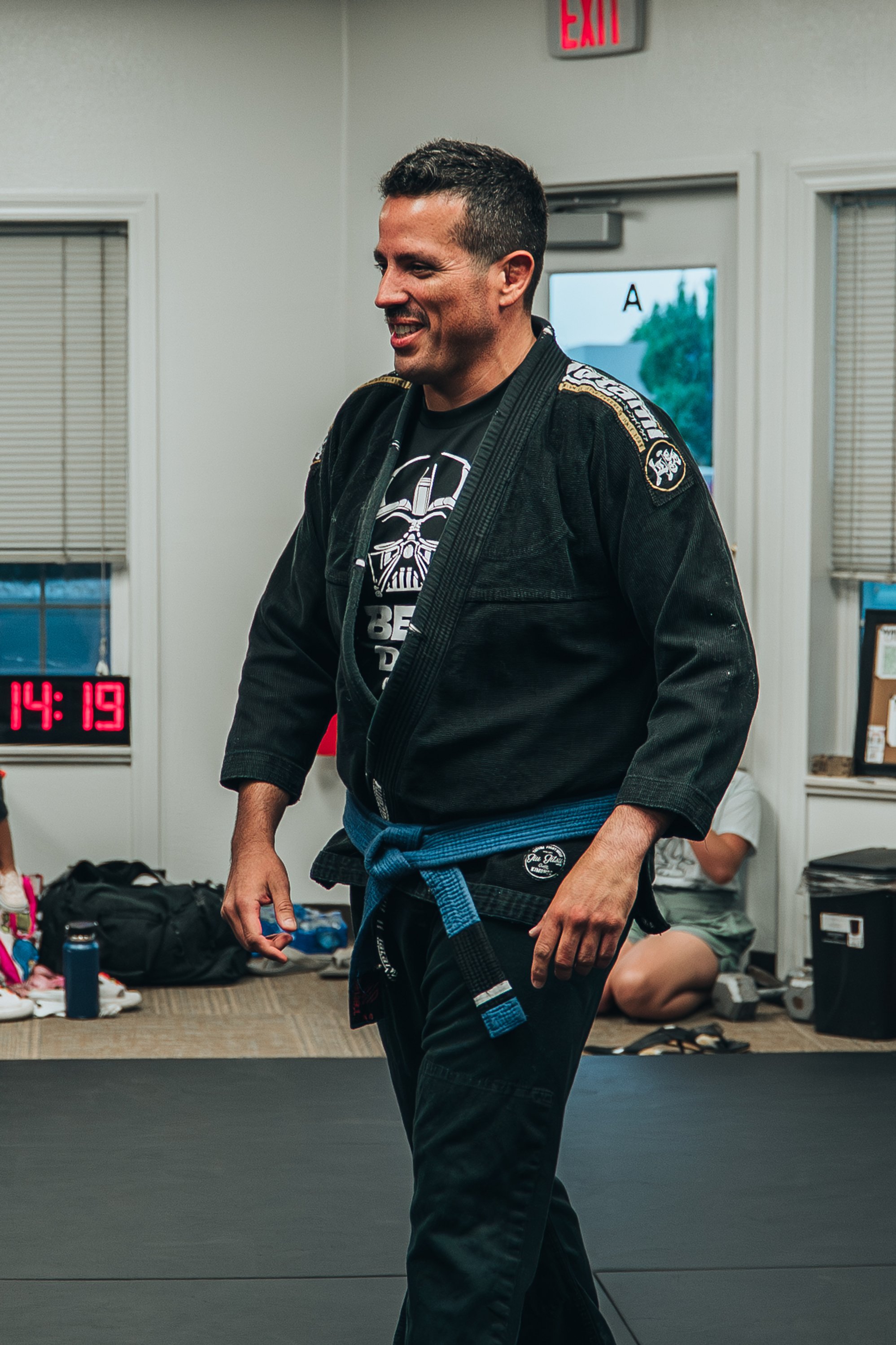 A man in a black Brazilian Jiu-Jitsu gi with a blue belt smiling in a martial arts gym.