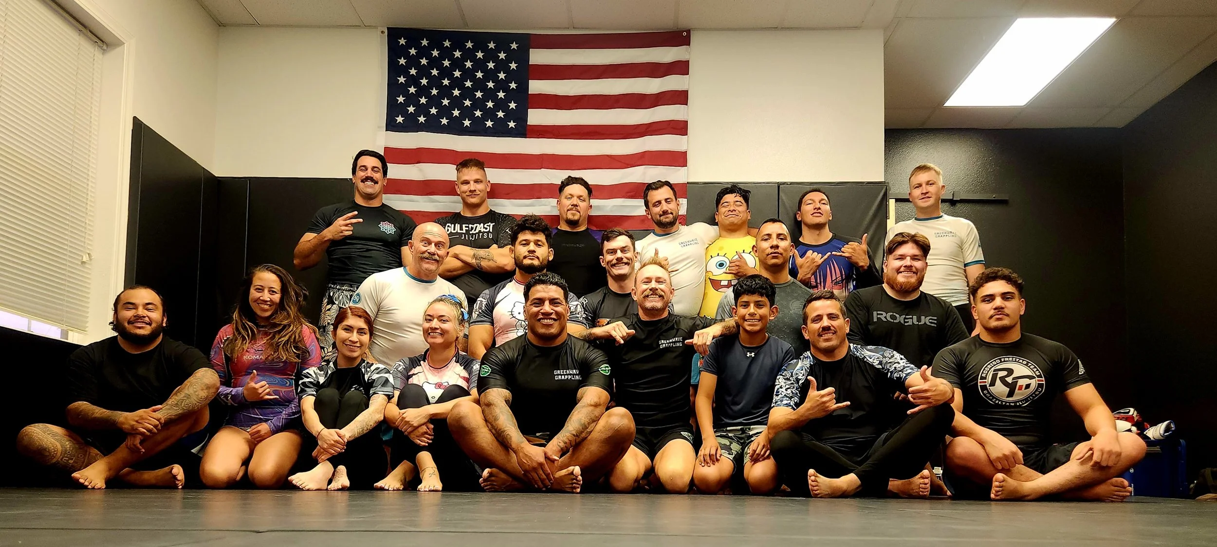 Group of people in a martial arts gym posing for a photo with an American flag in the background. Some are sitting cross-legged on the floor, others are standing behind them, making various hand gestures, and smiling.