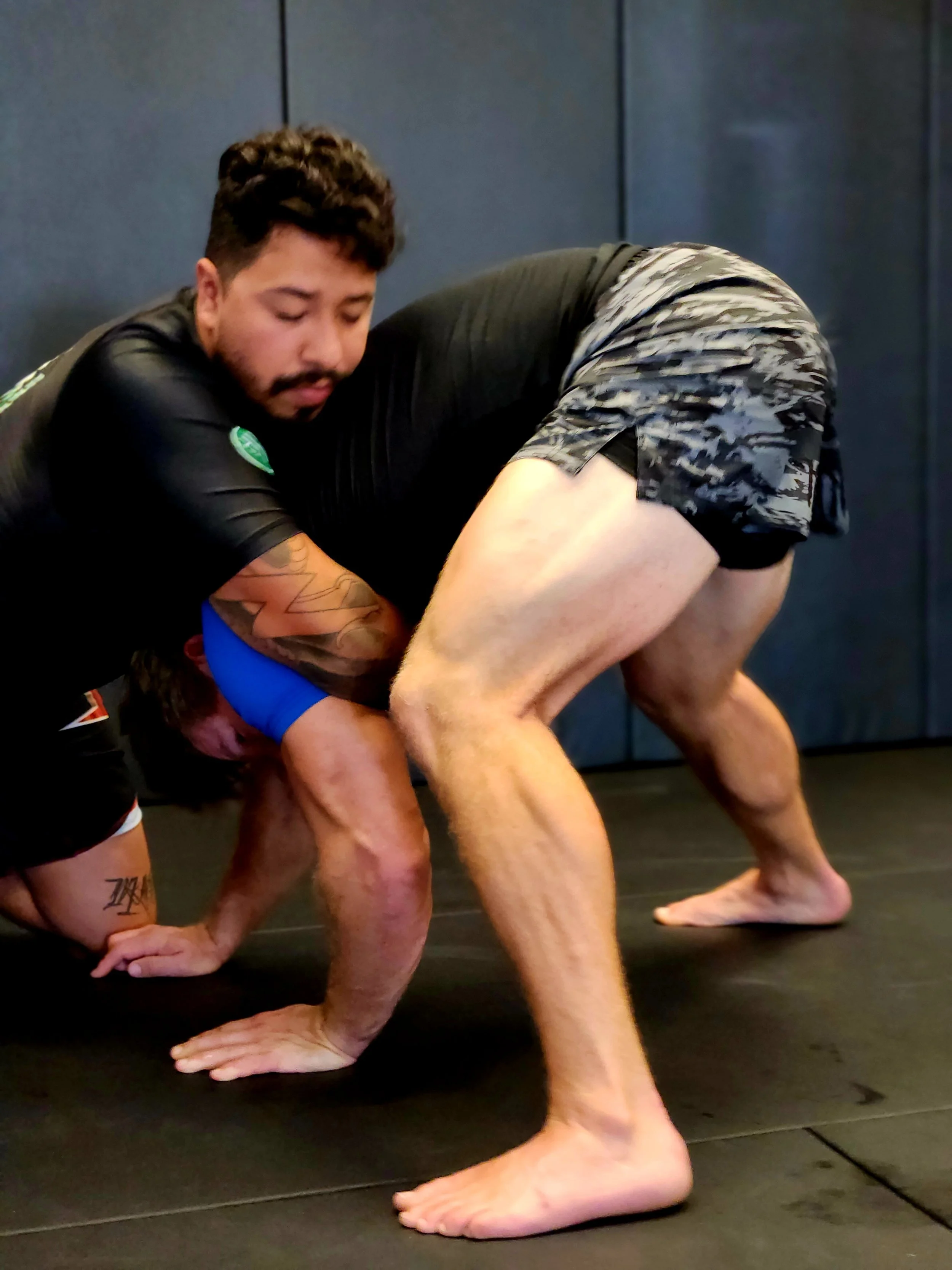 Two men practicing Brazilian Jiu-Jitsu on a black mat, one in a black rash guard and shorts, and the other in a blue rash guard and black shorts, engaged in grappling.