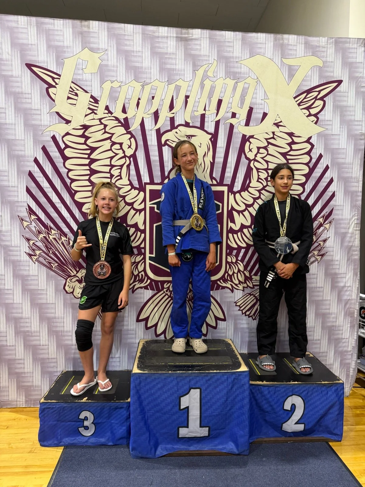 Three young girls standing on a winners' podium at a martial arts competition, with medals around their necks. The girl in the center, on the first-place platform, is wearing a blue gi. The girl on the right, on the second-place platform, is in a black gi. The girl on the left, on the third-place platform, is in a black gi with a knee brace. The backdrop features a large logo with a stylized eagle and the word 'TappologyX'.
