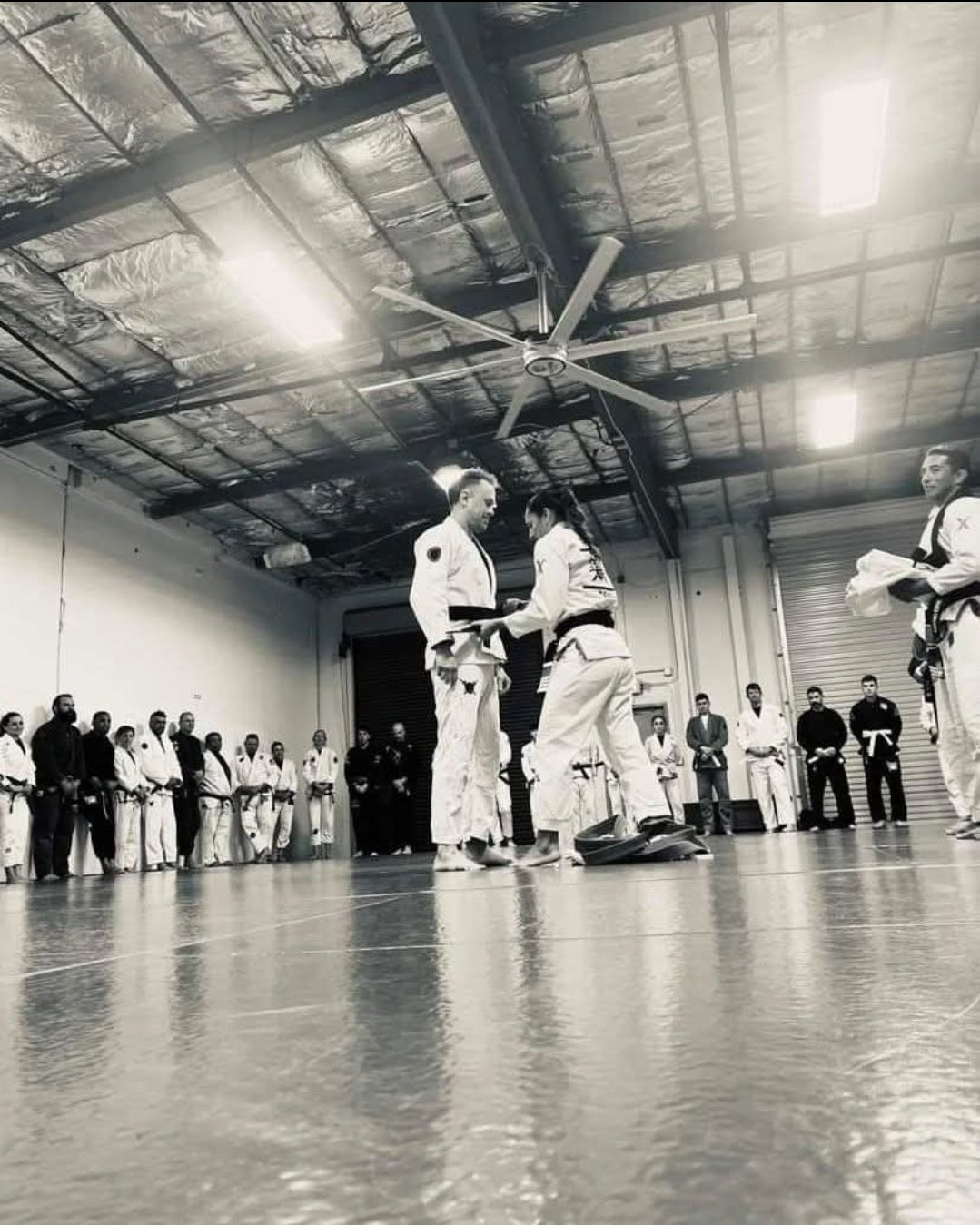 Brazilian Jiu-Jitsu practitioners in a training dojo, with two fighters in the center exchanging grips, surrounded by onlookers and other practitioners, in a black-and-white photo.