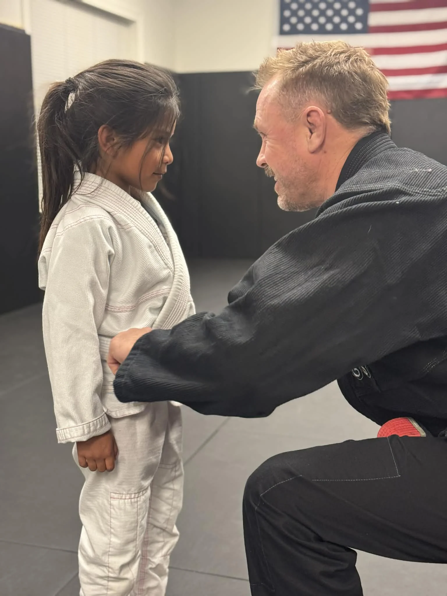 A young girl in a white martial arts uniform standing face to face with an adult male in a black gi, both smiling, in a martial arts dojo with an American flag in the background.