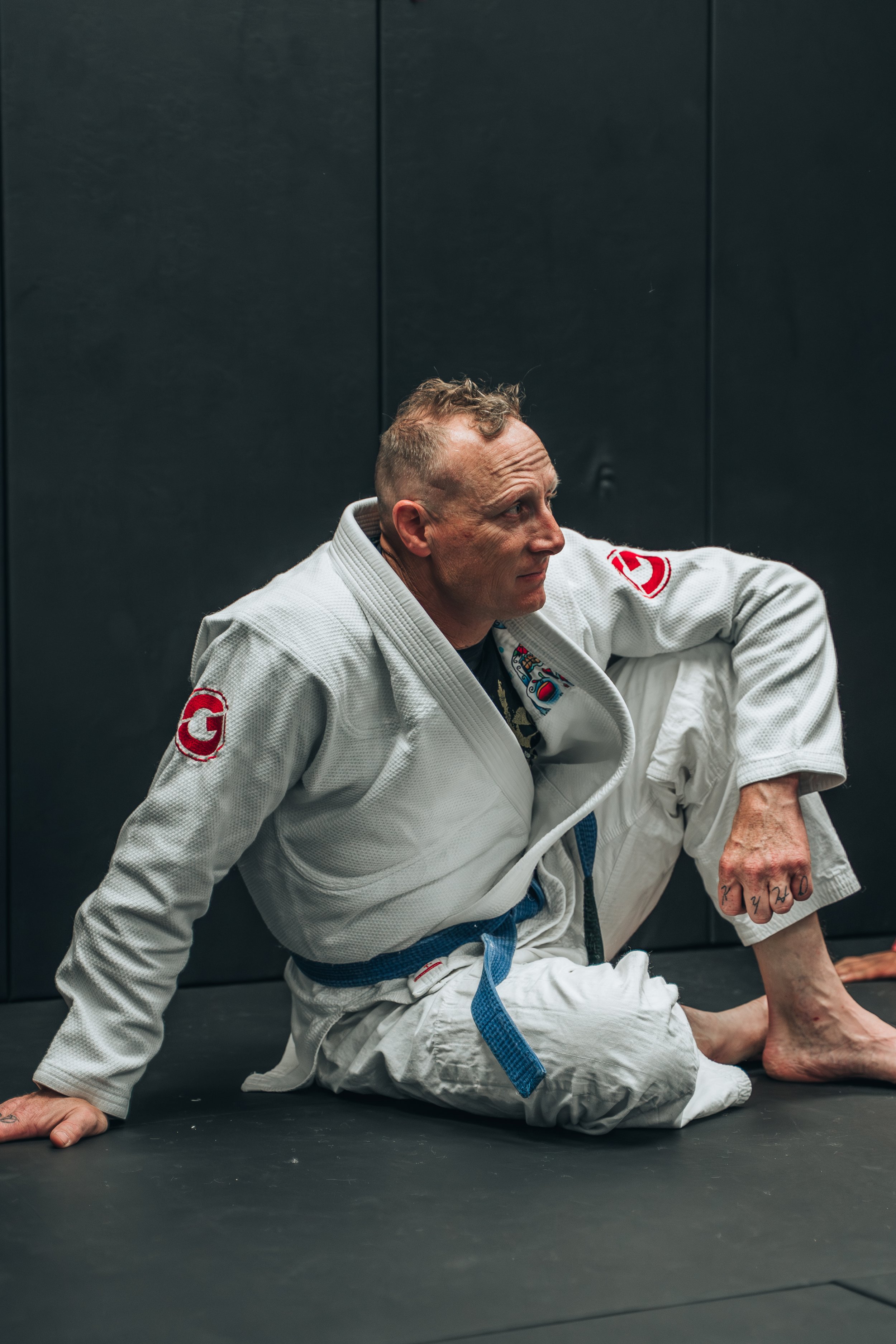 A male Brazilian Jiu-Jitsu practitioner wearing a white gi, with a blue belt, sitting on the ground in a grappling stance on a black mat in a training facility.