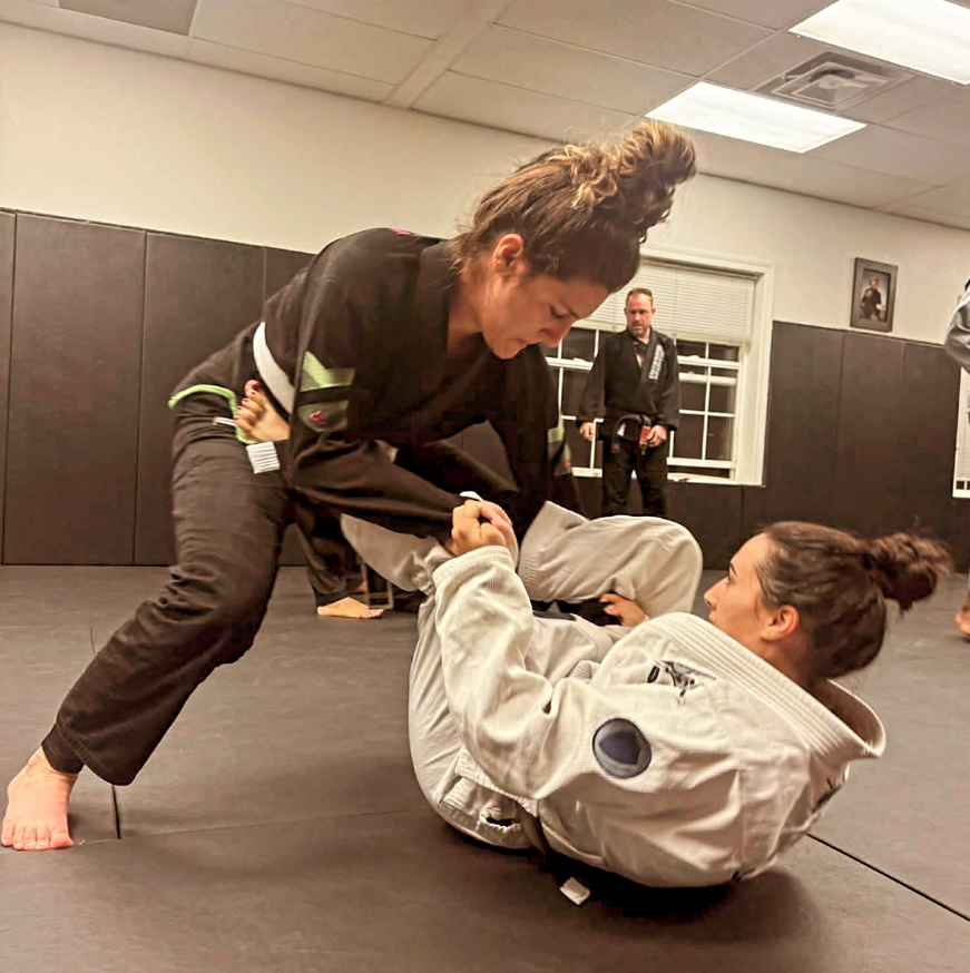Two women practicing Brazilian Jiu-Jitsu on a mat, one standing and holding the other's hands, on a training mat in a martial arts studio with other practitioners in the background.
