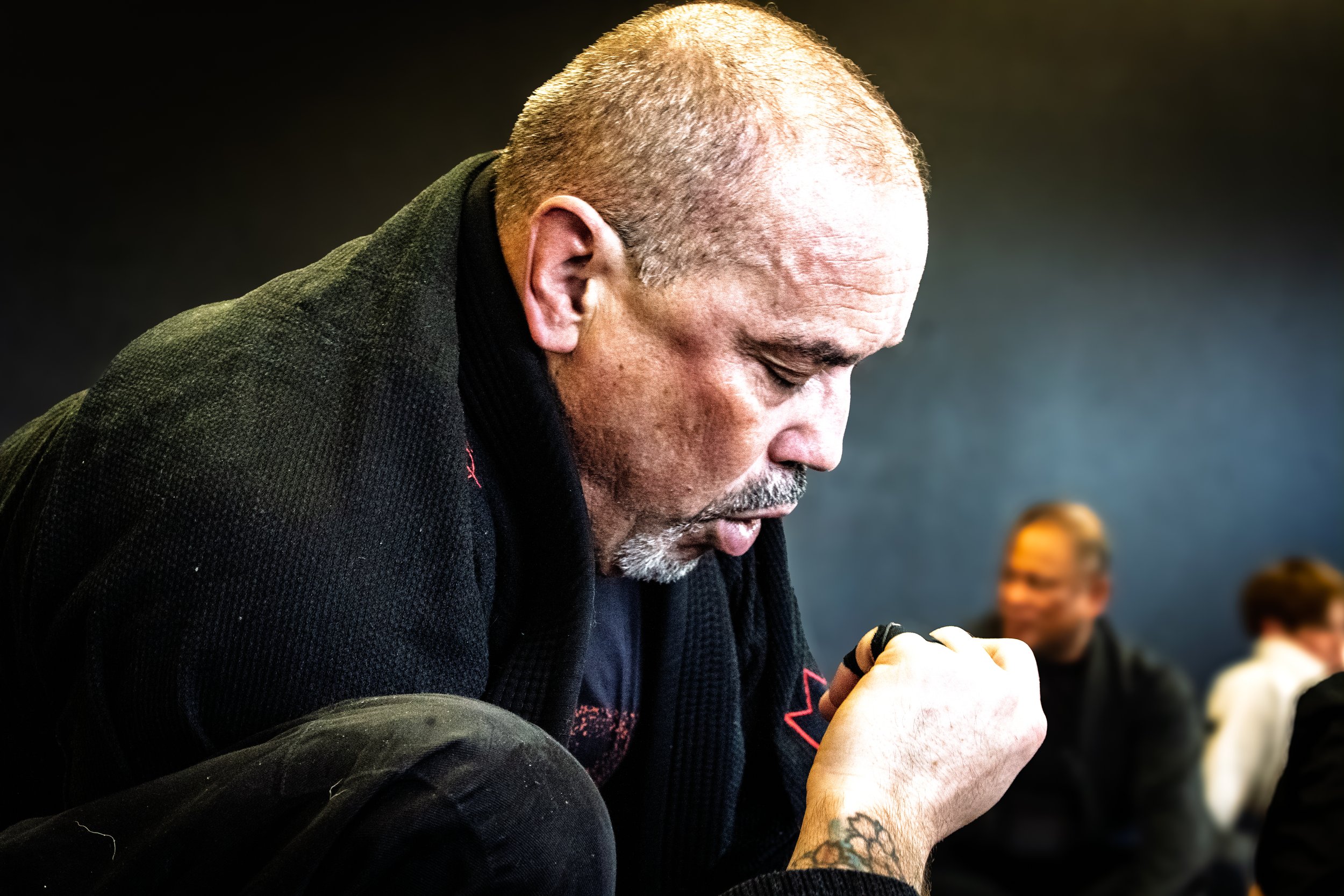 A man with short gray hair, a beard, and a tattooed hand is kneeling with his head bowed, eyes closed, in a moment of prayer or reflection. He is wearing a black jacket, and there are blurred people in the background.