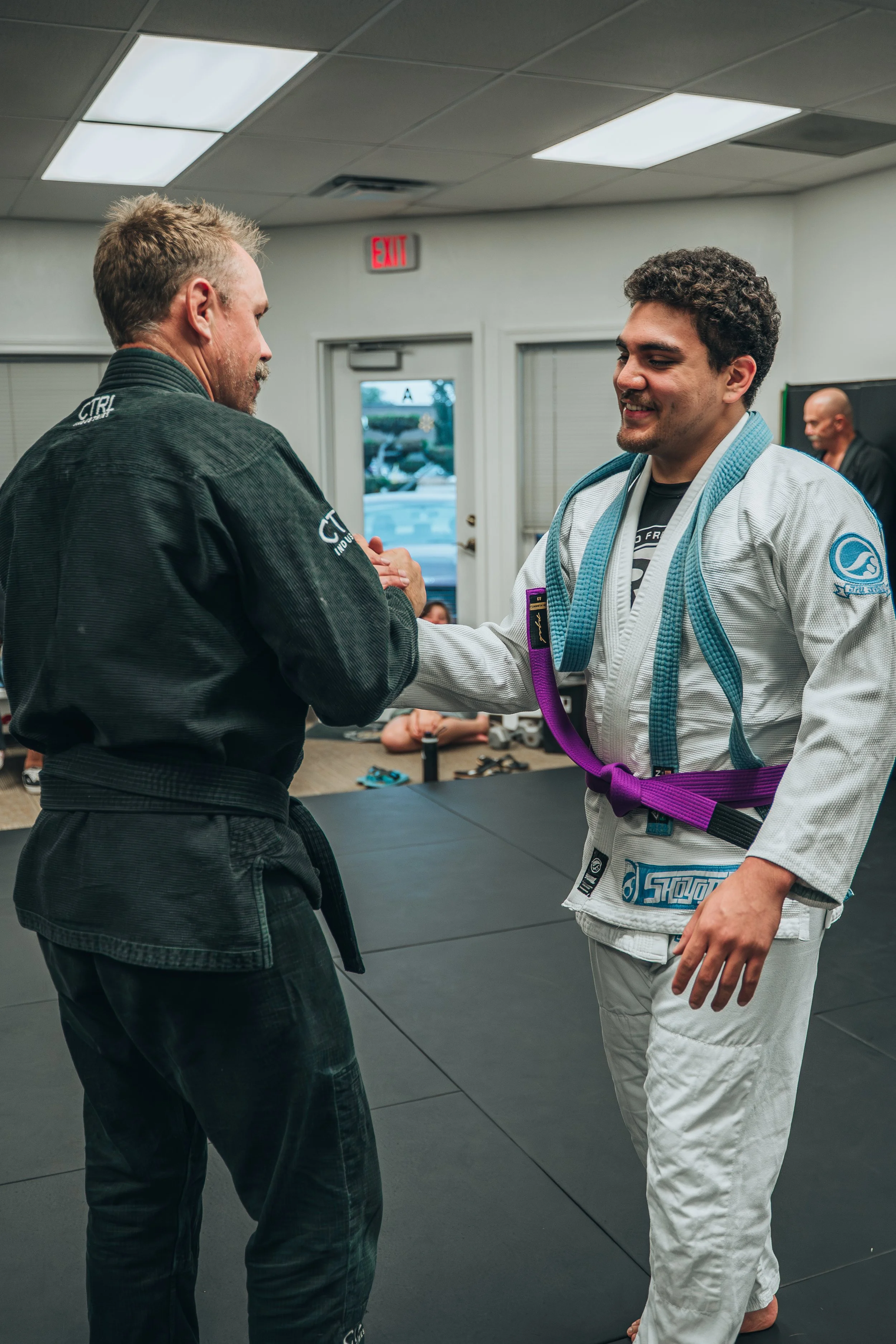 Two men shaking hands in a Brazilian Jiu-Jitsu training facility, one in a black gi and the other in a white gi with a purple belt, smiling.