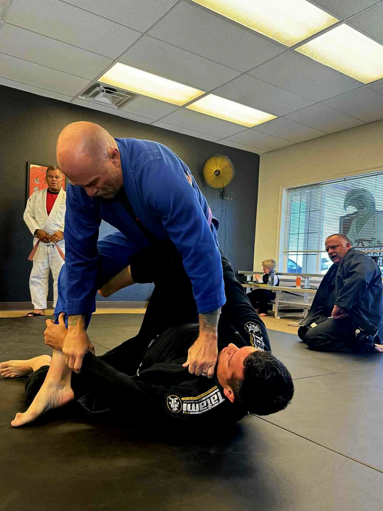 Two men practicing Brazilian Jiu-Jitsu on a mat. One man in a black gi is on the ground, while the other man in a blue gi is on top. A man in a white gi with a red shirt stands in the background observing. Another man, also in a black gi, is kneeling near the window, and a woman is seated at a table.