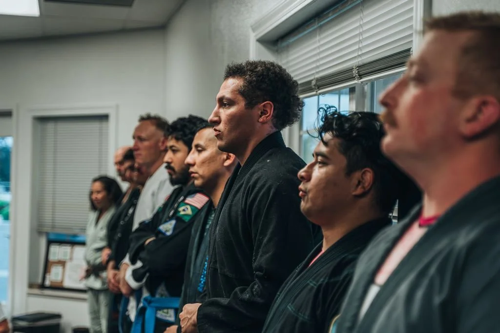 Group of people in martial arts uniforms standing in a line, facing sideways, indoors near windows with blinds.