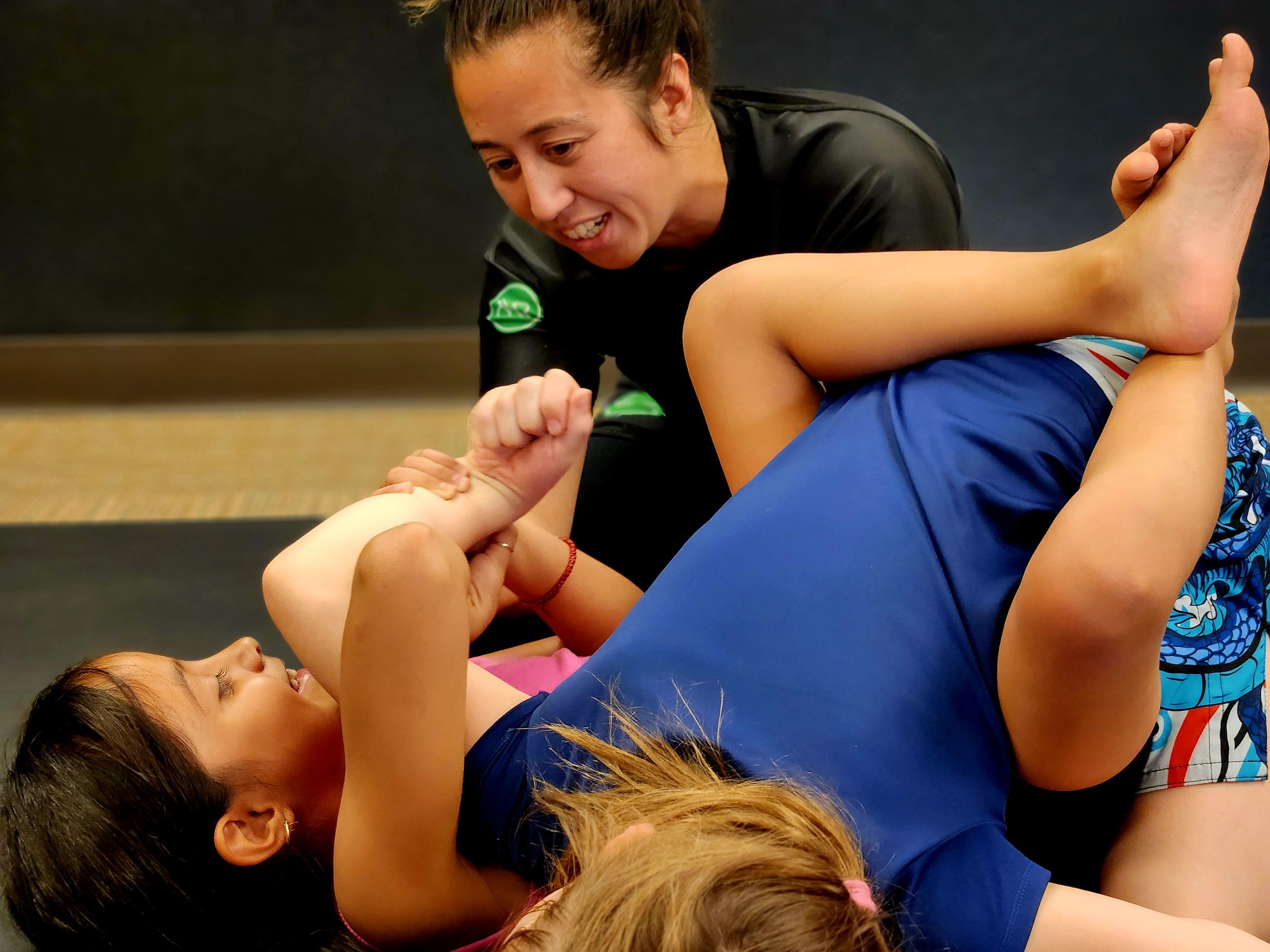 Two young girls practicing Brazilian Jiu-Jitsu on a mat with an instructor assisting them.