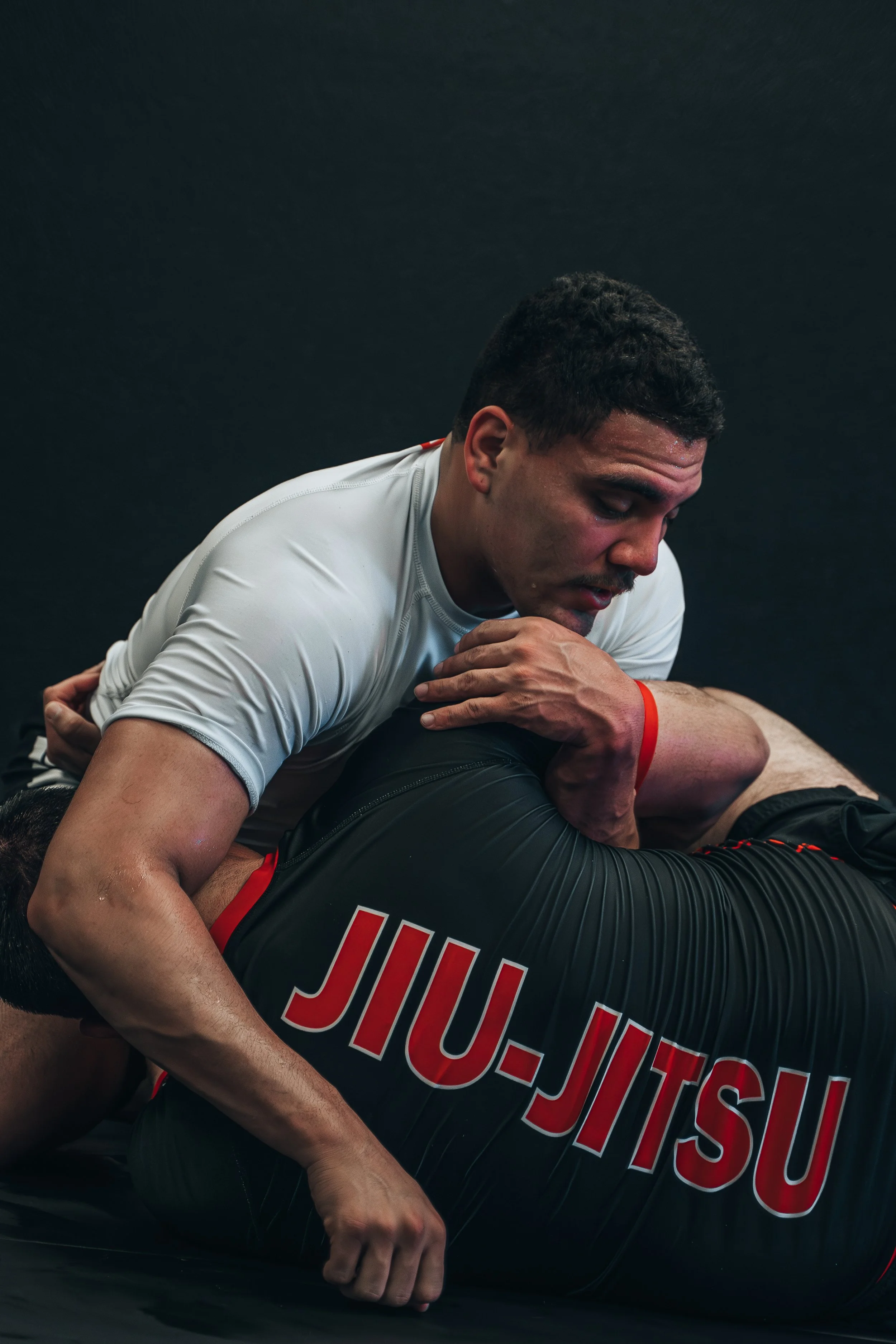 Two men practicing Brazilian Jiu-Jitsu on black mats, one in a white shirt and the other in a black rash guard with red lettering.
