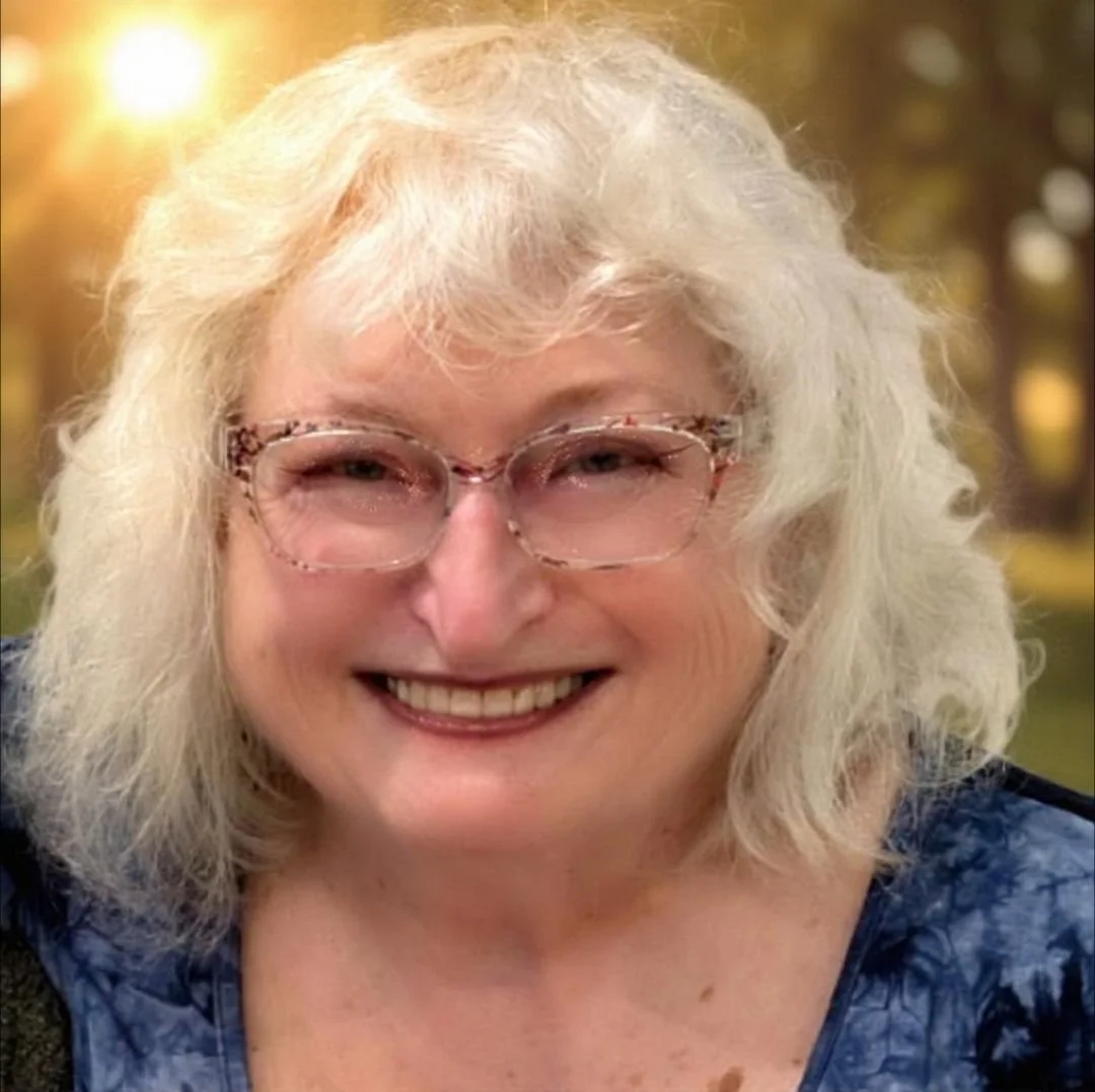 A smiling older woman with curly white hair and glasses, outdoors in a sunny setting with trees.