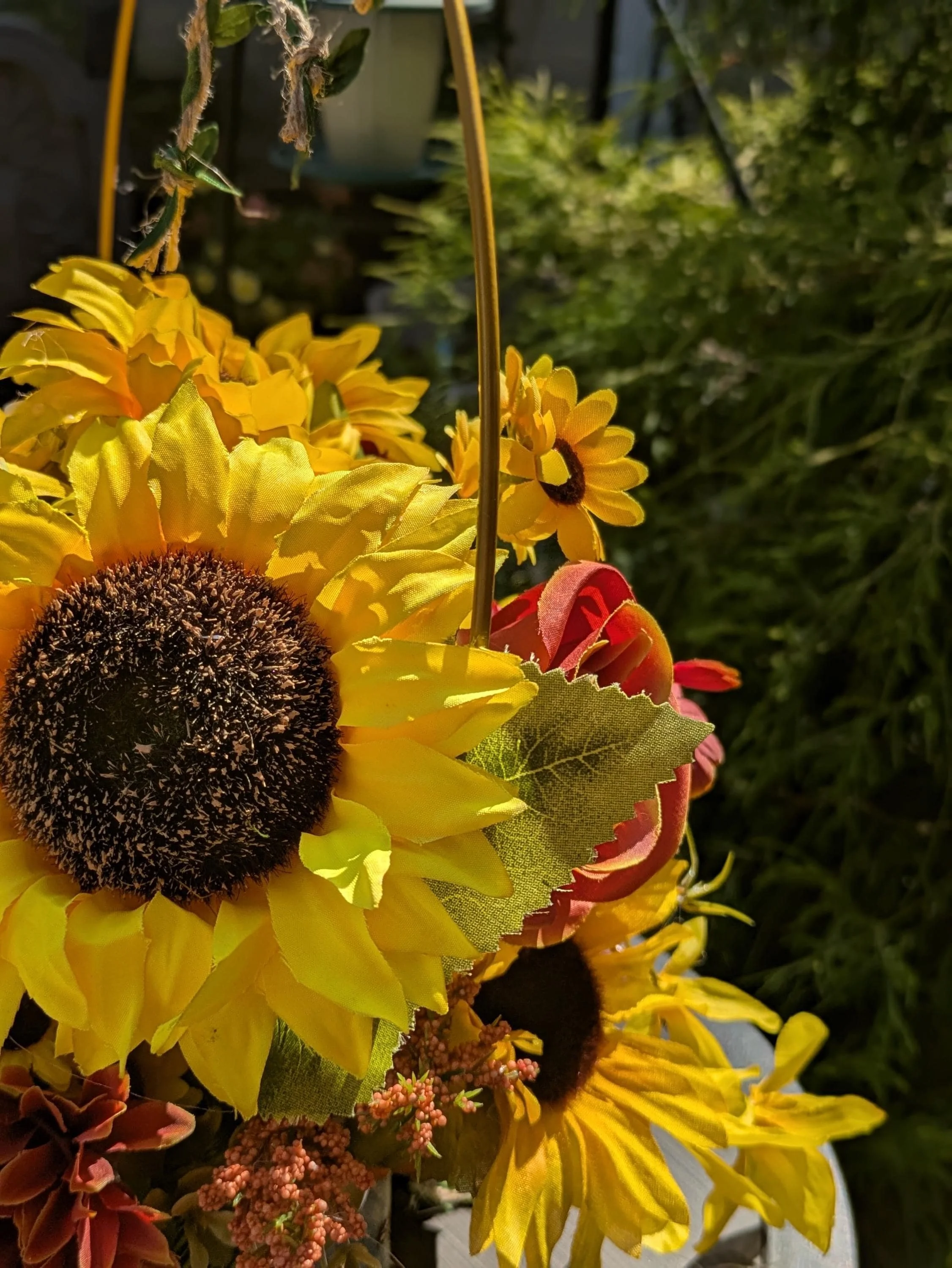 Close-up of a hanging flower arrangement outdoors, featuring large yellow sunflowers, small daisies, and artificial green leaves, illuminated by sunlight with a blurred garden background.