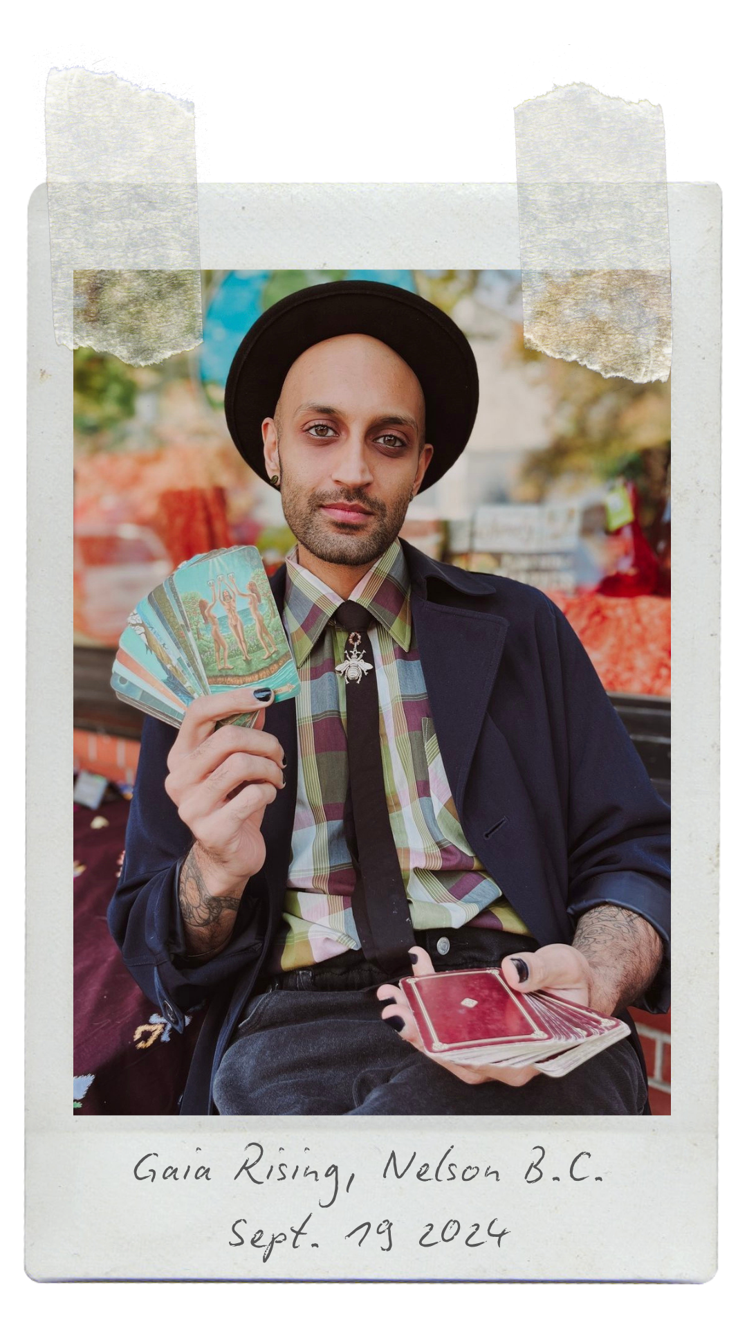 A person with a bald head and wearing a black hat and dark jacket, sitting outdoors surrounded by market stalls, holding tarot cards in one hand and fanning a deck of red-backed cards with the other. The photo has a Polaroid-style border with handwritten caption: 'Gaia Rising, Nelson B.C. Sept. 19 2024'.