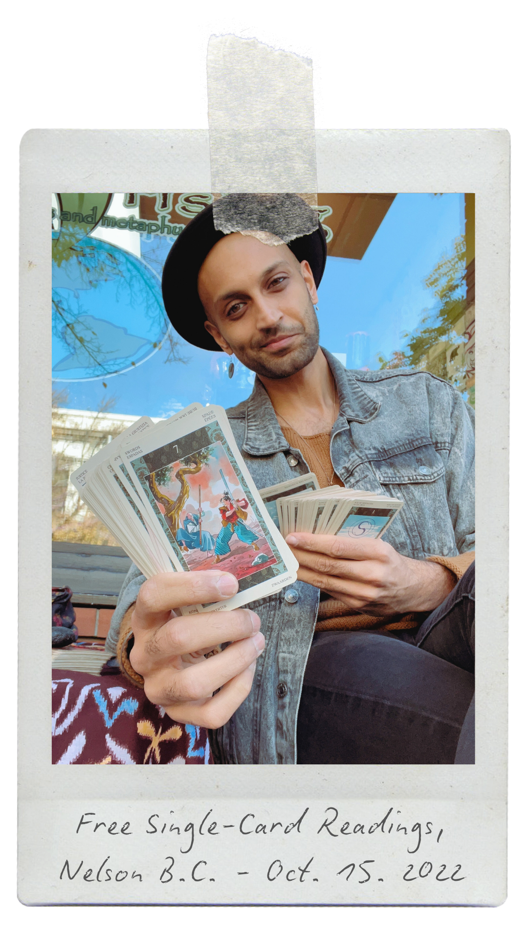 A man wearing a black hat and denim jacket holding tarot cards outdoors with a sign in the background. Handwritten note at the bottom says, "Free Single-Card Readings, Nelson B.C. - Oct. 15, 2022."