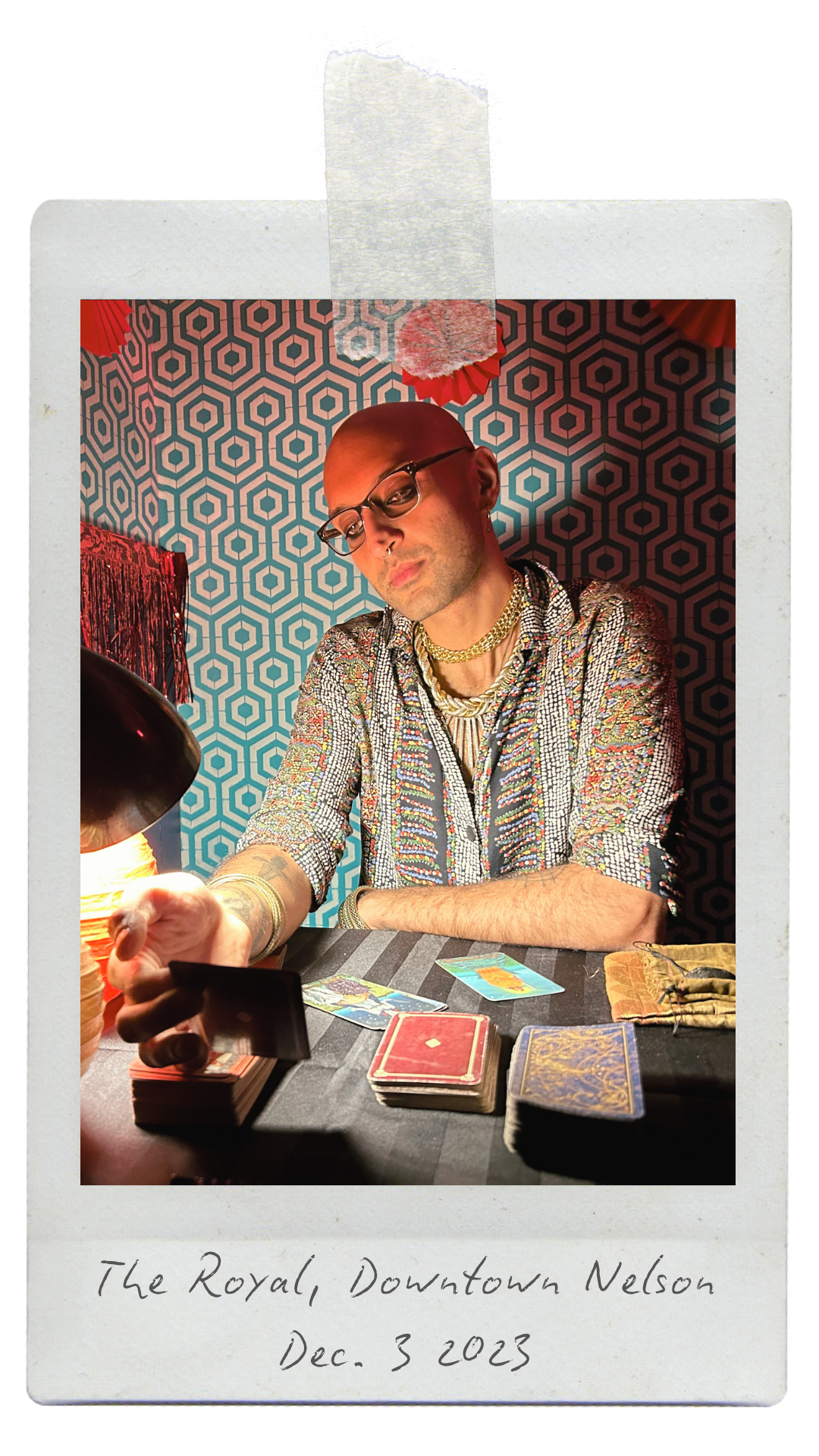 A person with glasses and jewelry sitting at a table with tarot cards, under decorative lighting and a patterned backdrop, with notes that say 'The Royal, Downtown Nelson Dec. 3 2023'.