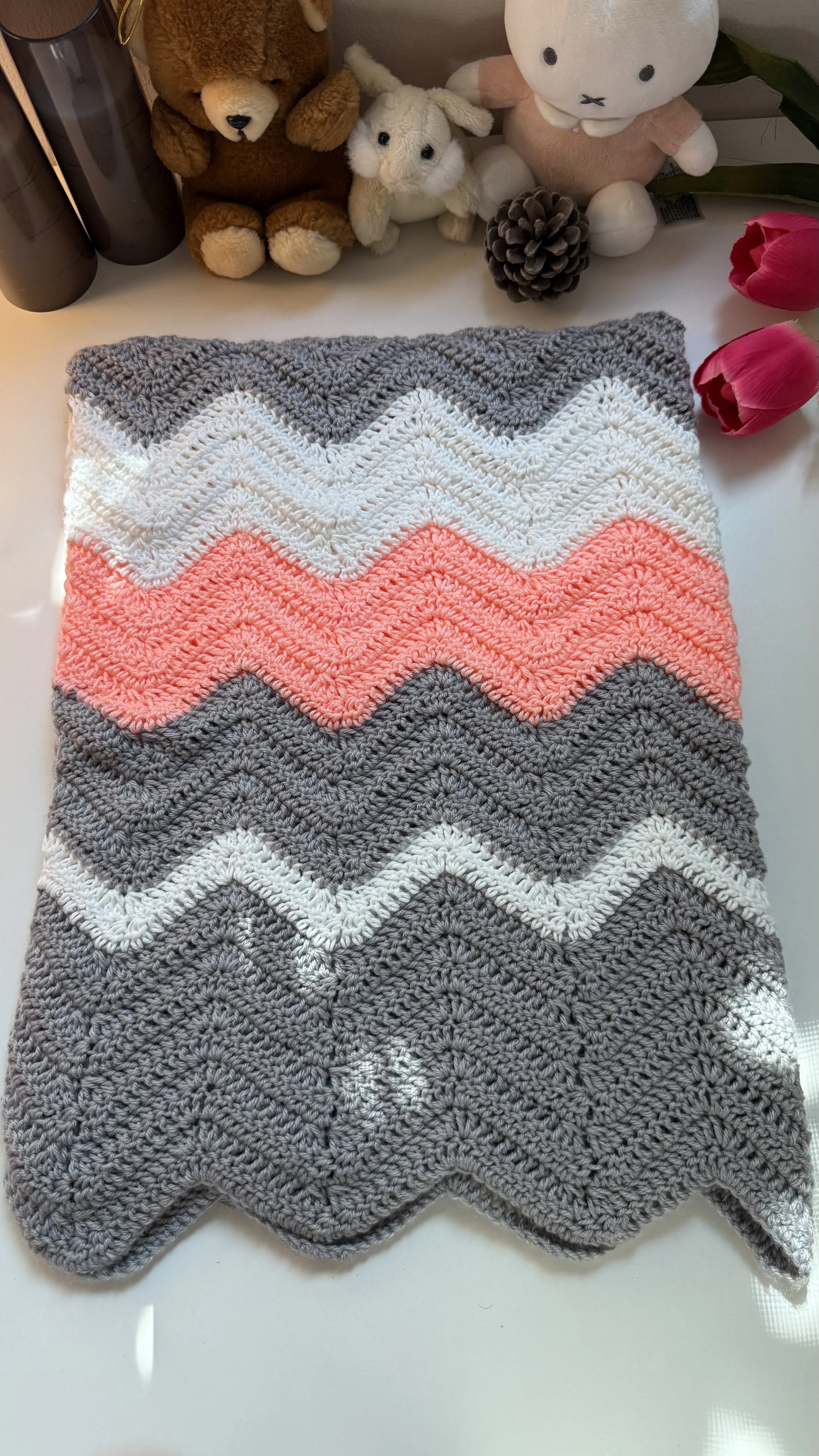 Close-up top view of a handmade crochet baby blanket featuring a textured zigzag chevron ripple pattern with alternating stripes of charcoal grey, white, cream, and vibrant pastel coral pink.