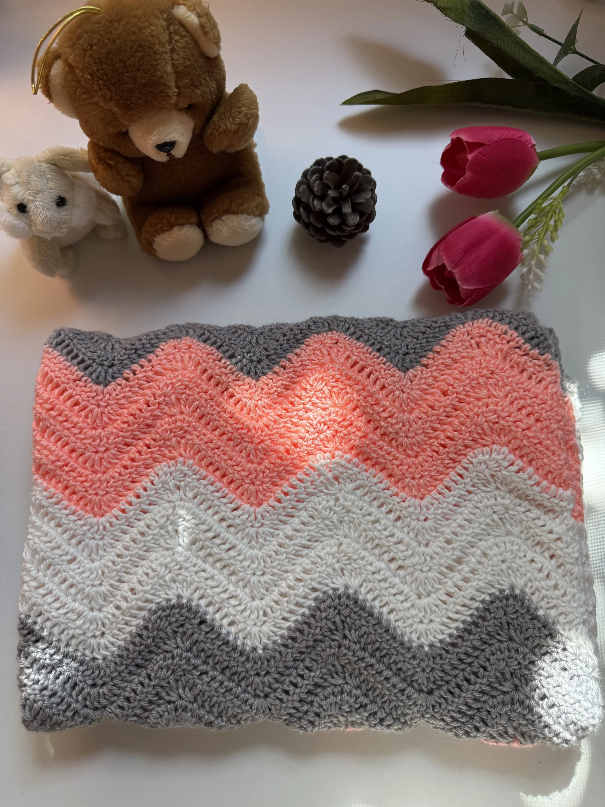 Close-up top view of a handmade crochet baby blanket featuring a textured zigzag chevron ripple pattern with alternating stripes of charcoal grey, white, cream, and vibrant pastel coral pink.