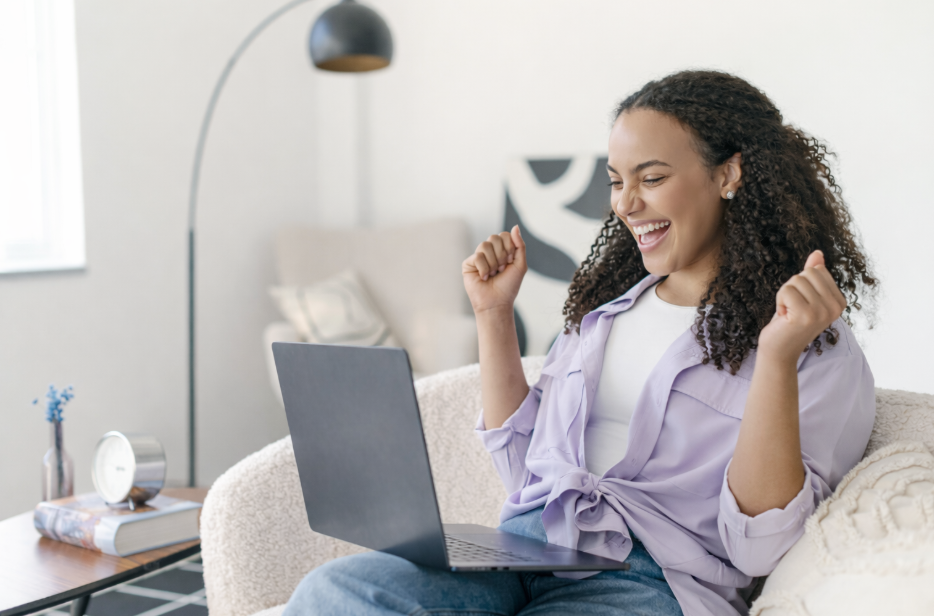 A woman celebrating excitedly while sitting on a beige couch, using a silver laptop in a bright living room with a white wall, a black floor lamp, and a side table with a clock, a book, and a small vase with blue flowers
