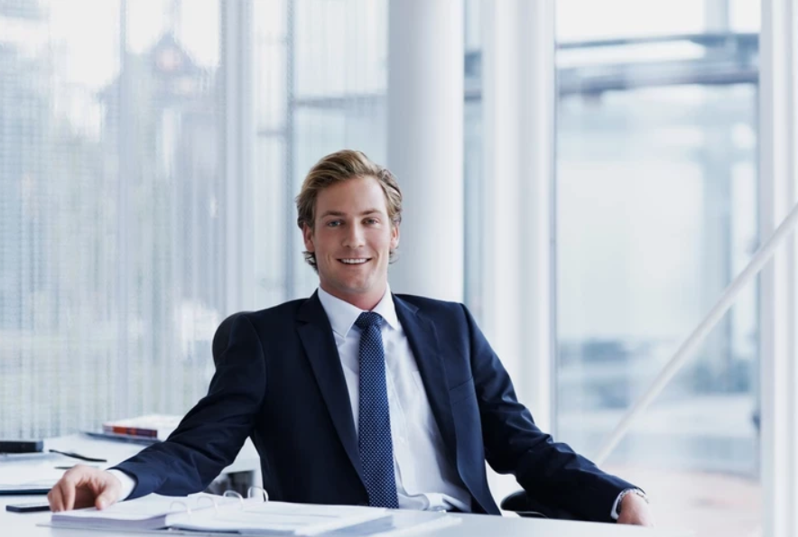 Businessman sitting at a desk in a modern office with large windows, smiling at the camera.