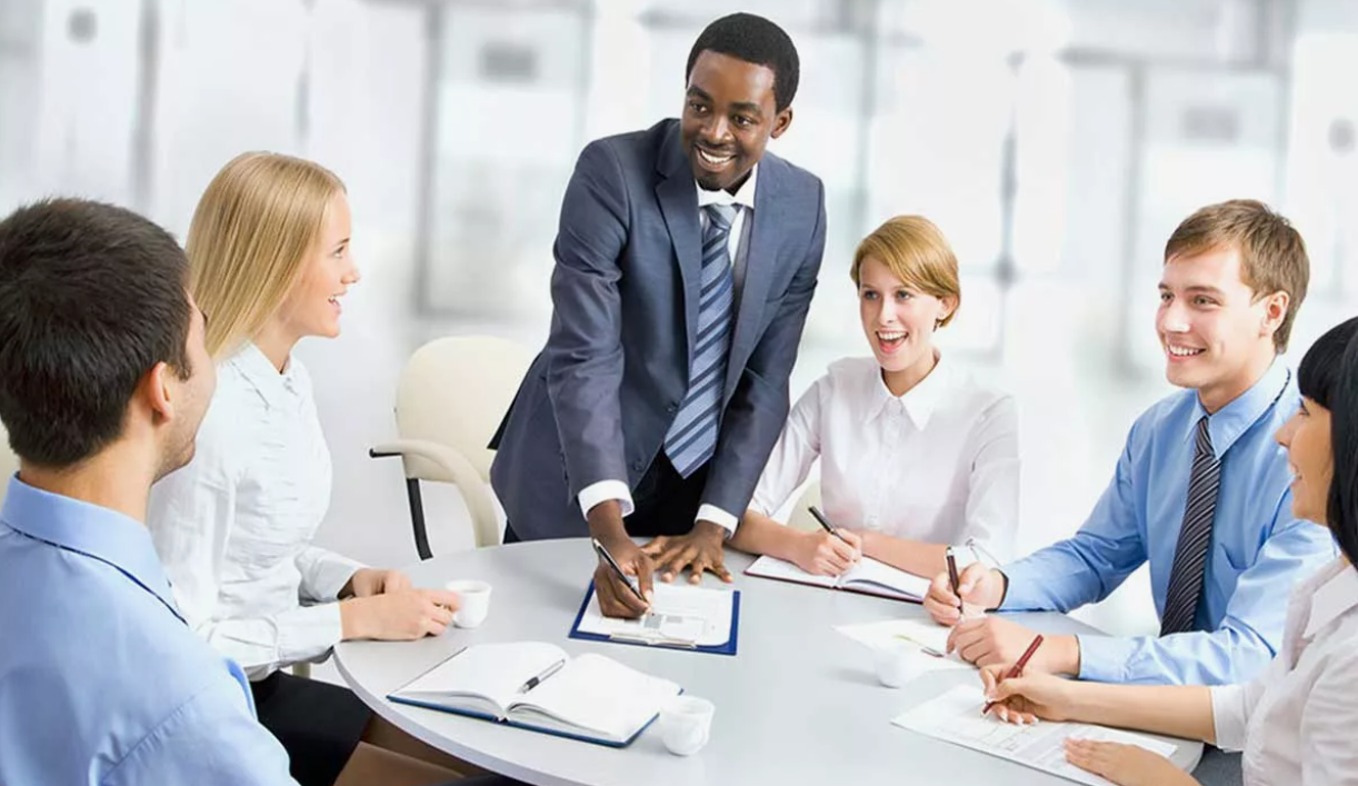 A diverse group of six people in business attire sitting around a conference table in a modern office, engaging in a discussion with a smiling man standing and writing on a clipboard.