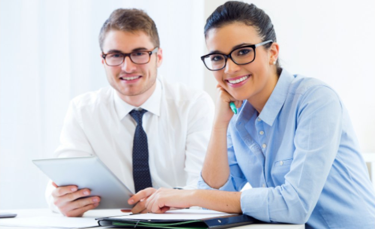 Two professional people, a man and a woman, sitting at a table with documents, smiling at the camera in an office.