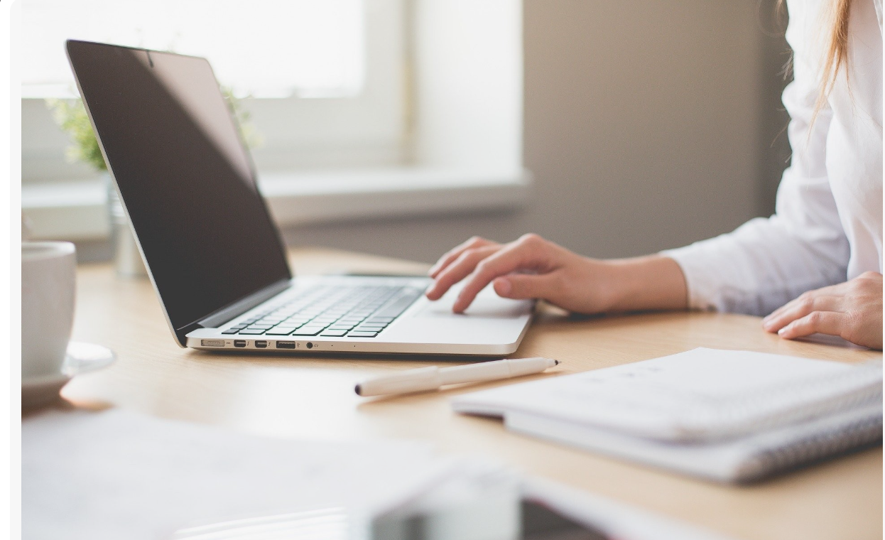 Person working on a silver laptop to learn English at a wooden desk with notebooks, a pen, and a cup nearby, near a window.