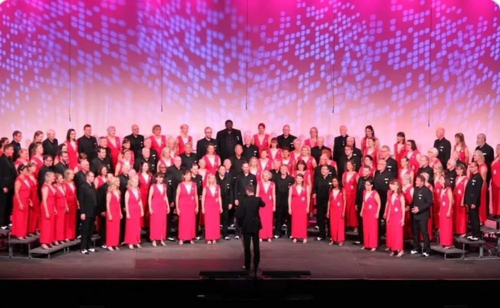 A large choir group dressed in red and black standing on risers on a stage, performing with a conductor in front of them, illuminated by a colorful light display.