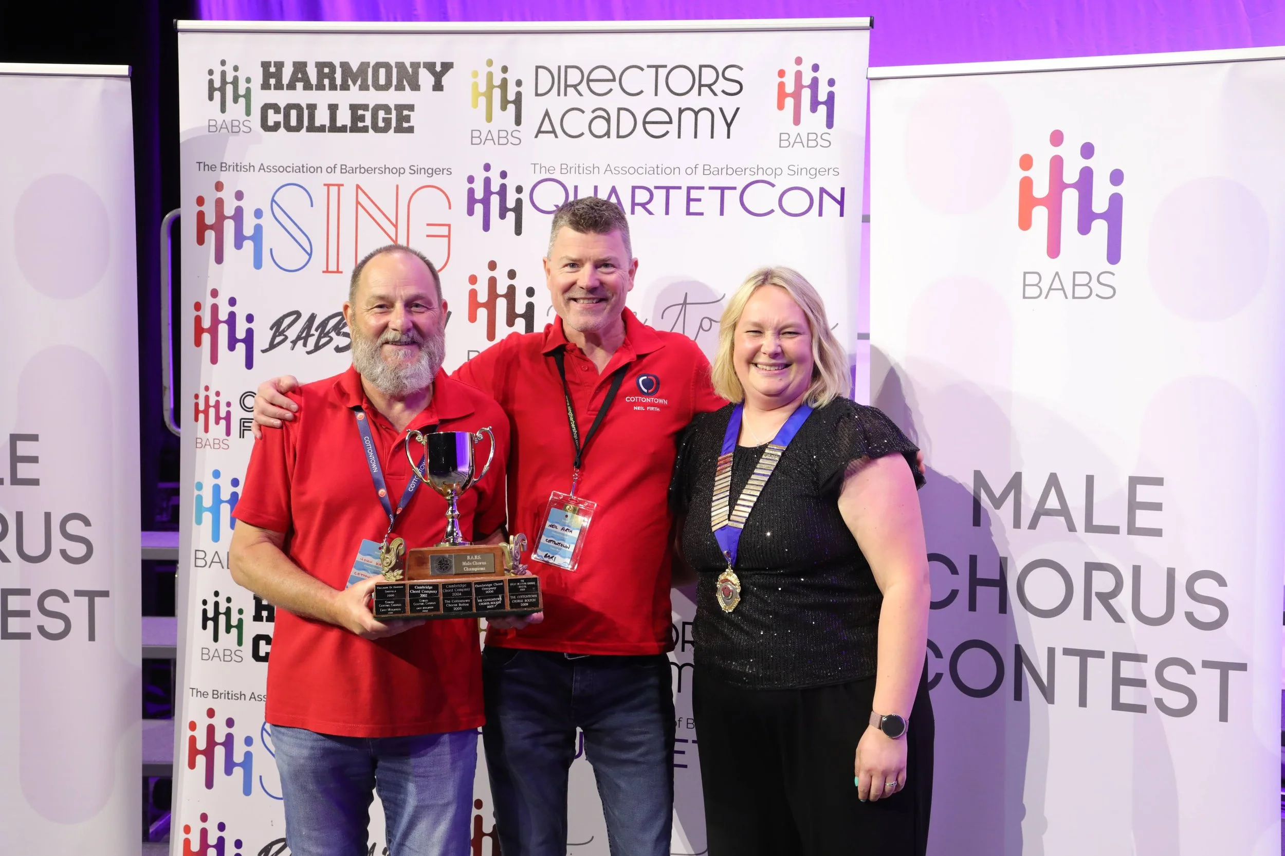 Three smiling individuals, two men and one woman, stand together at an event, with the man on the left holding a trophy. They are wearing red and black shirts, and all have medals around their necks. The backdrop features multiple banners with logos 