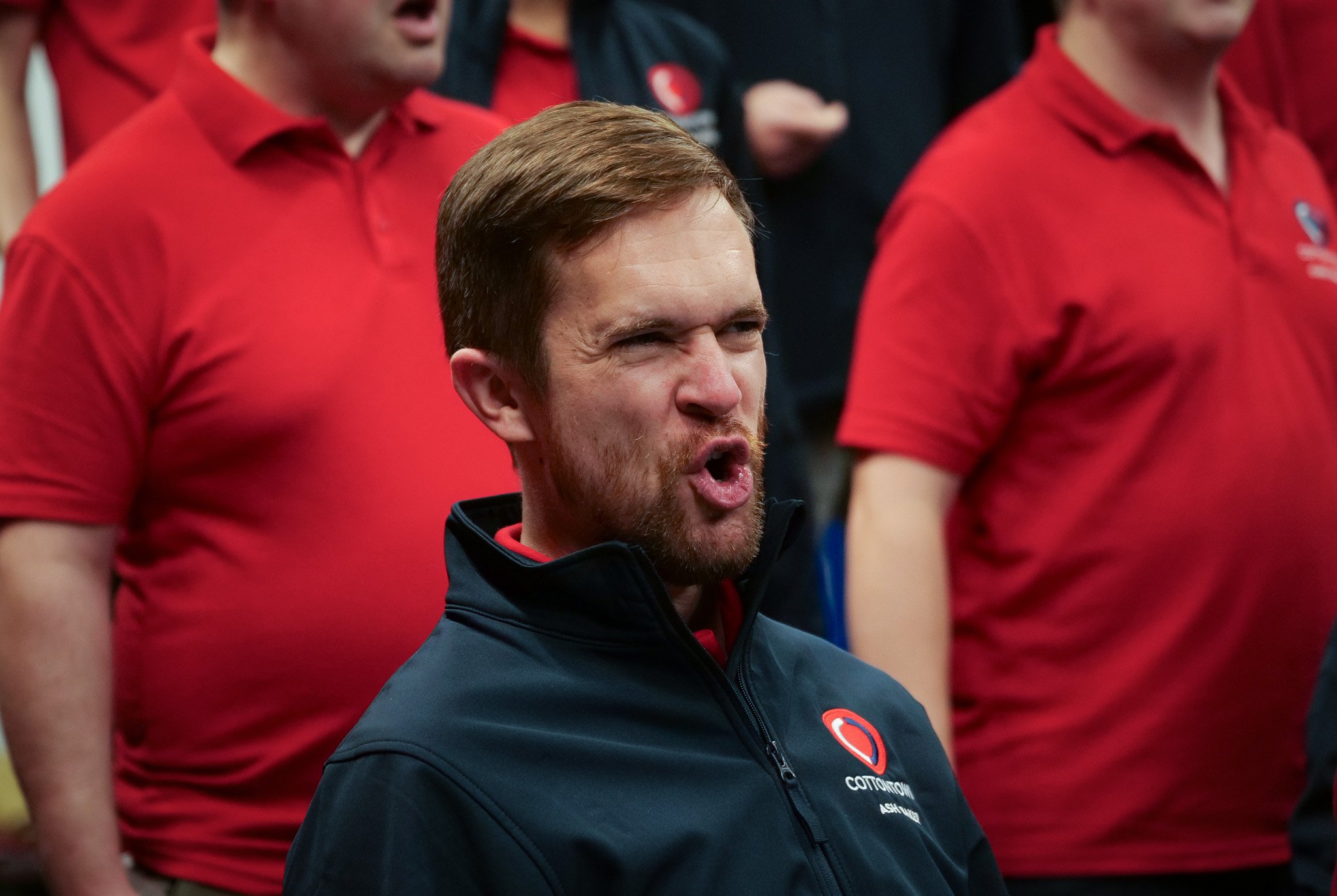 A man with a beard and short brown hair making a fierce face, surrounded by people in red shirts, at a crowd event.