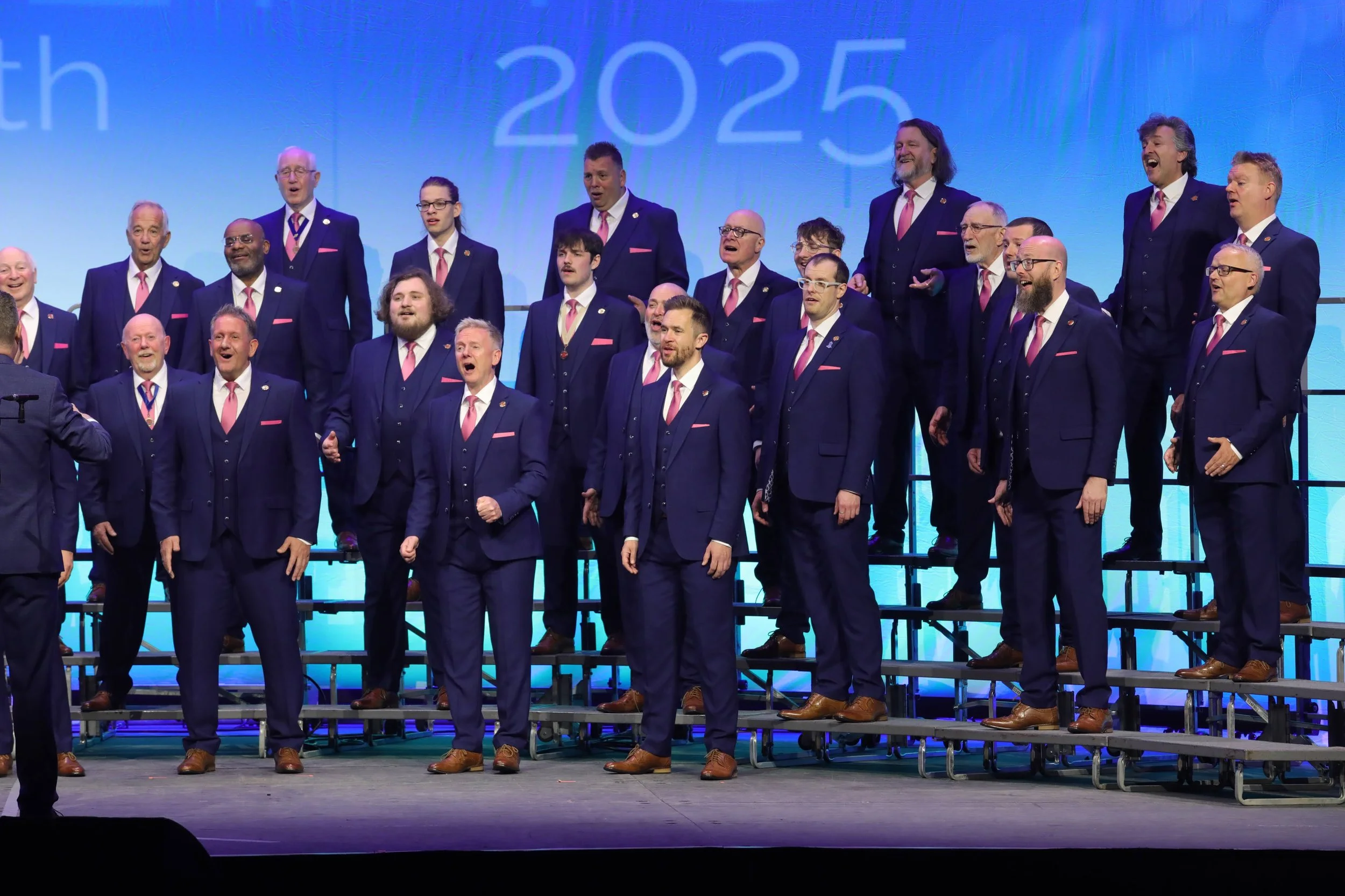 A men's choir dressed in navy suits with pink ties and pocket squares performing on a stage, standing on risers, with a blue background displaying '2025'.