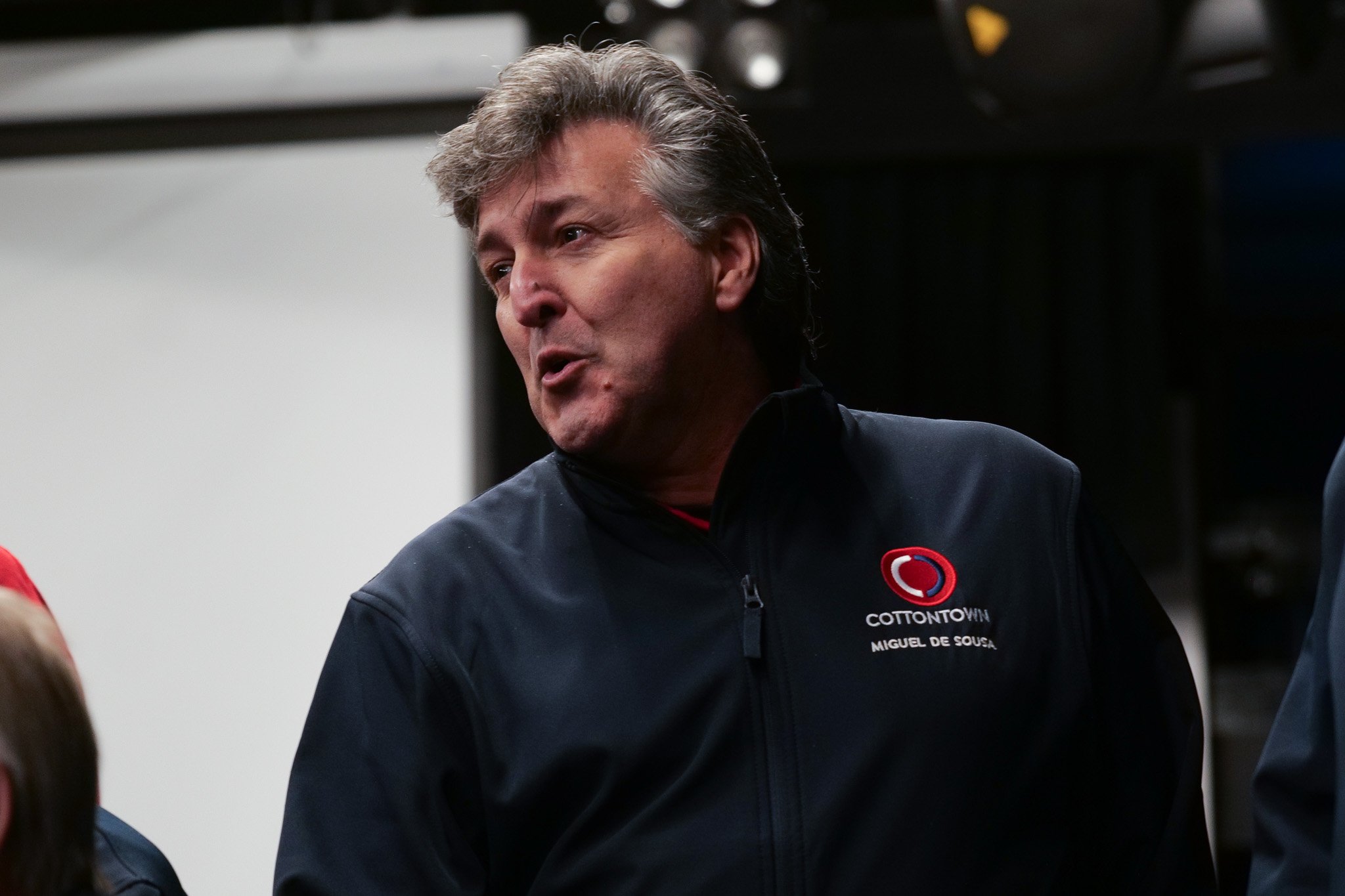 A middle-aged man with gray hair wearing a black jacket with a logo and text, speaking and looking to the side.