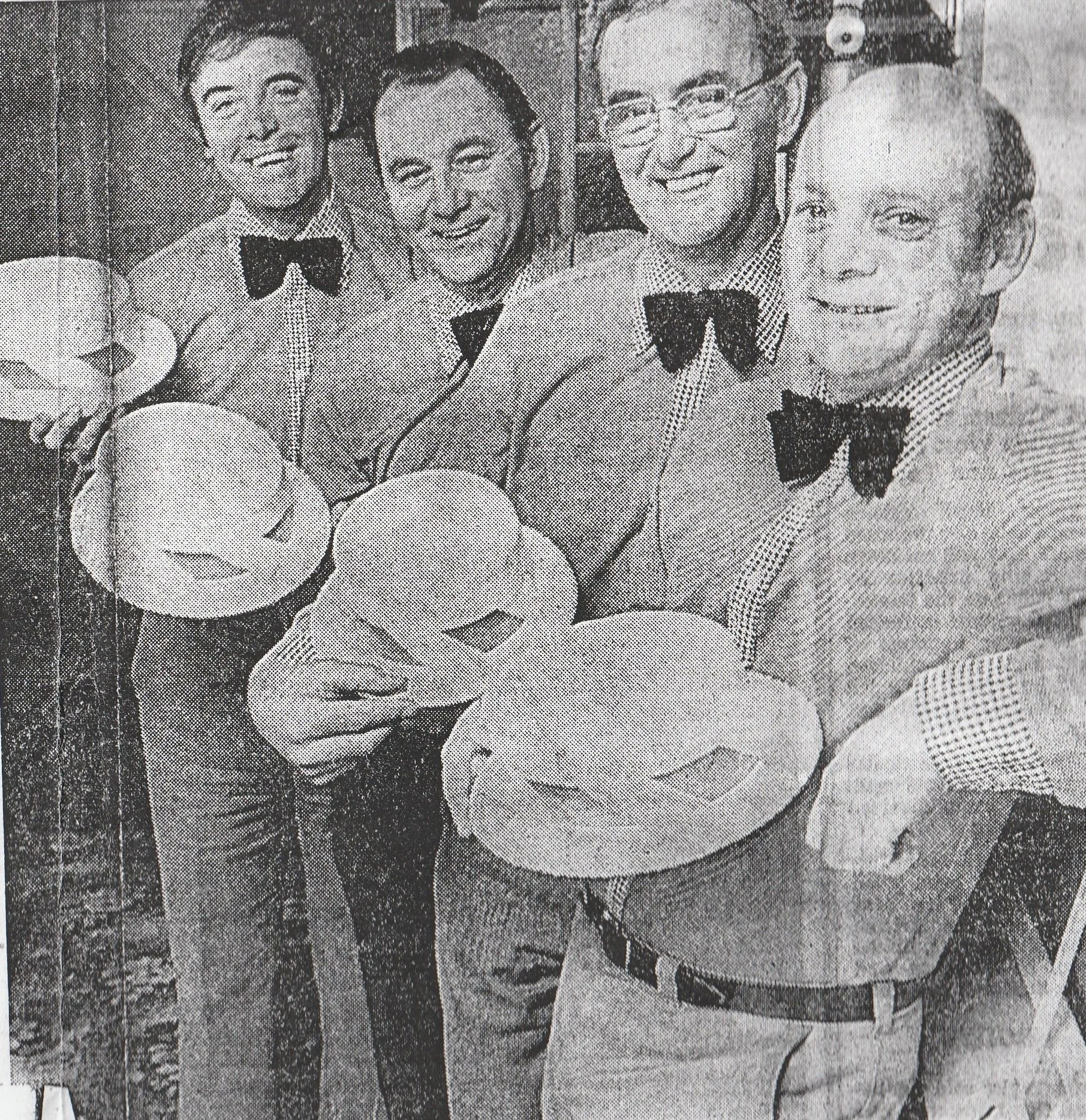 Four men dressed in suits with bow ties holding plates with boaters, smiling at the camera.